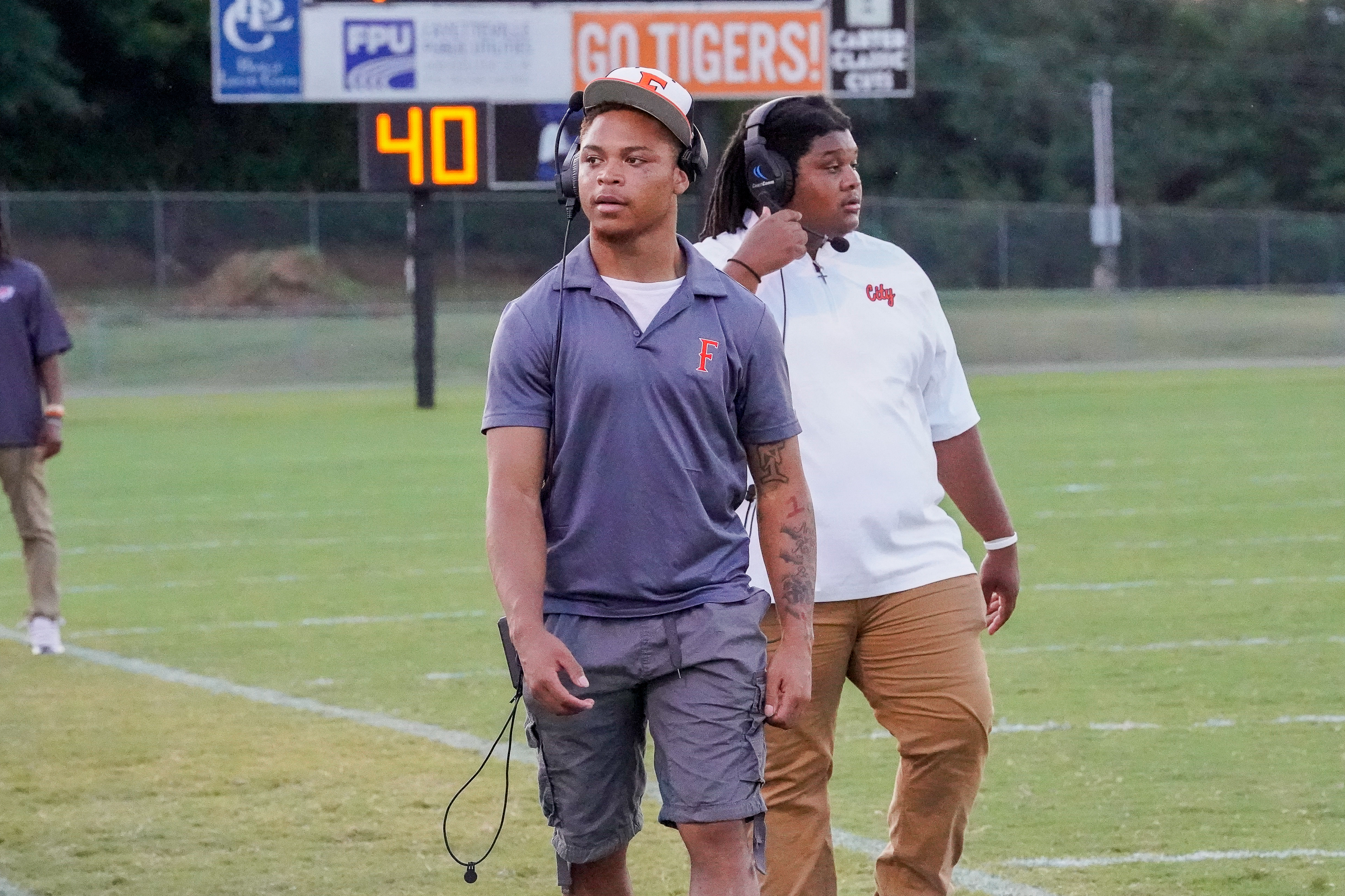 Two young men in athletic wear stand on a football field, with a scoreboard displaying "40" and "GO TIGERS!" in the background.