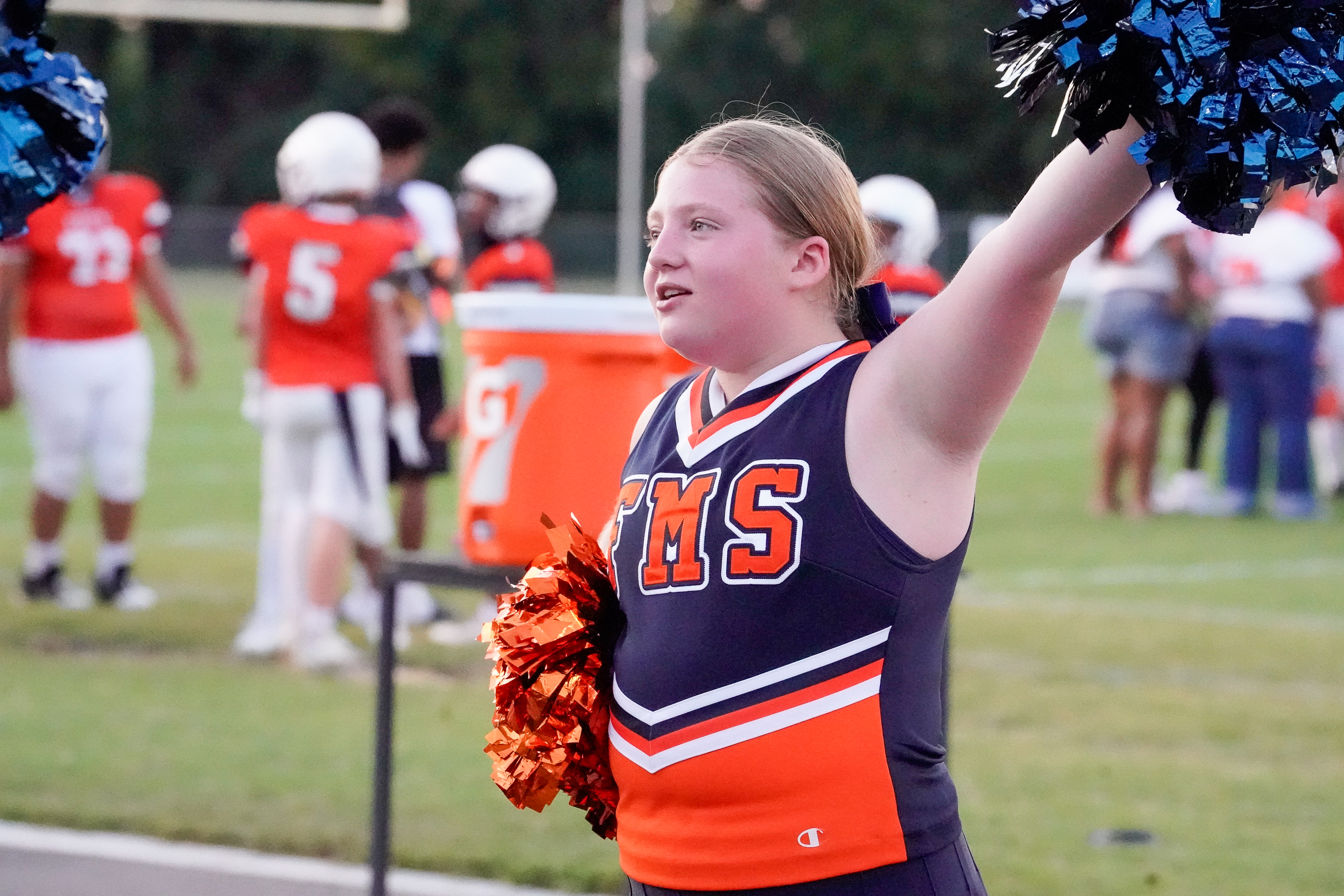 A young cheerleader with orange and blue pom-poms stands on a grassy field in the foreground, while blurred figures in football uniforms are visible in the background.