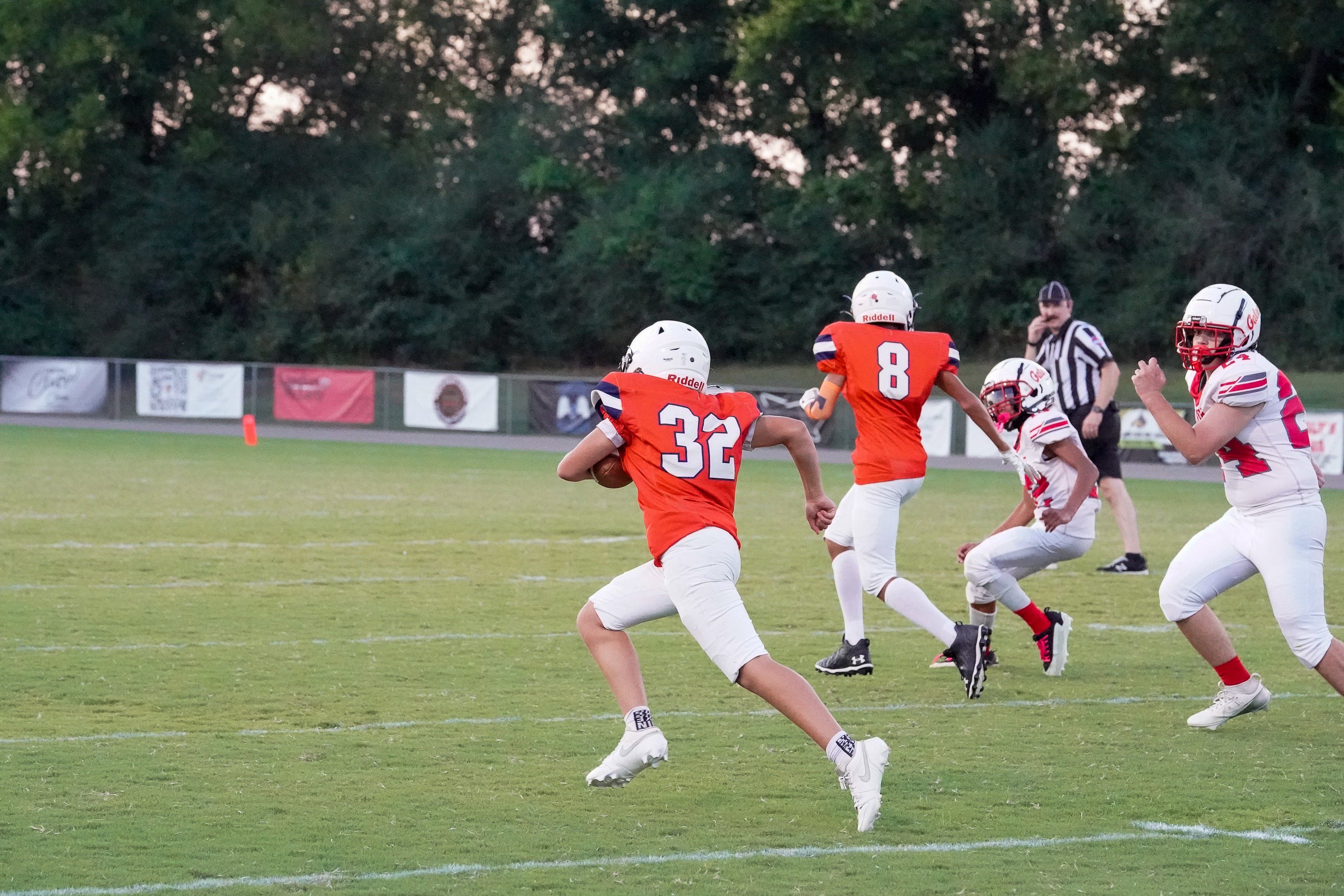 A football player in an orange jersey with the number 32 runs with the ball across a grassy field, while other players and a referee are visible in the background against a backdrop of trees and a stadium fence.