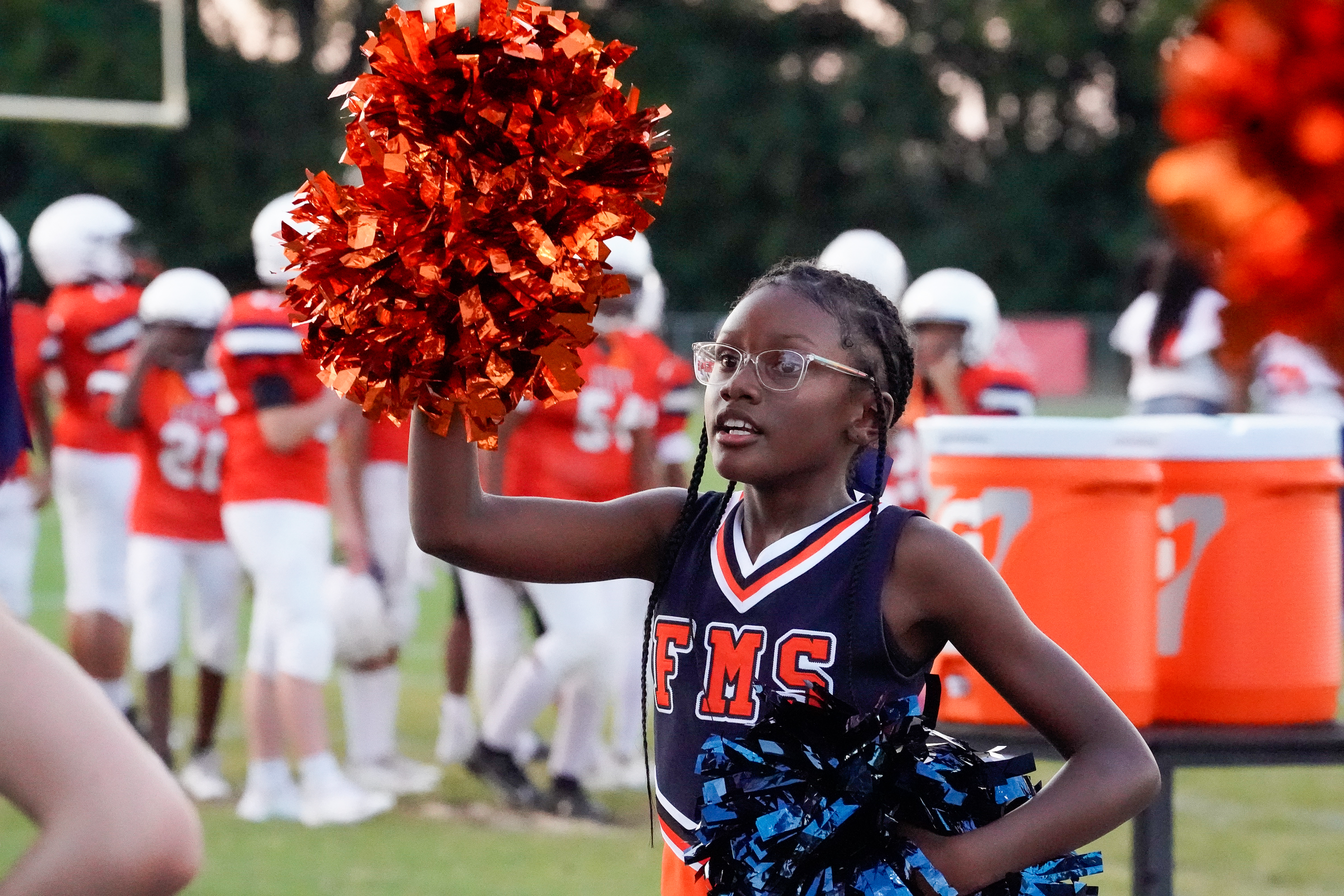 A young cheerleader with glasses and orange pom-poms in the foreground cheers for a football team in the background.