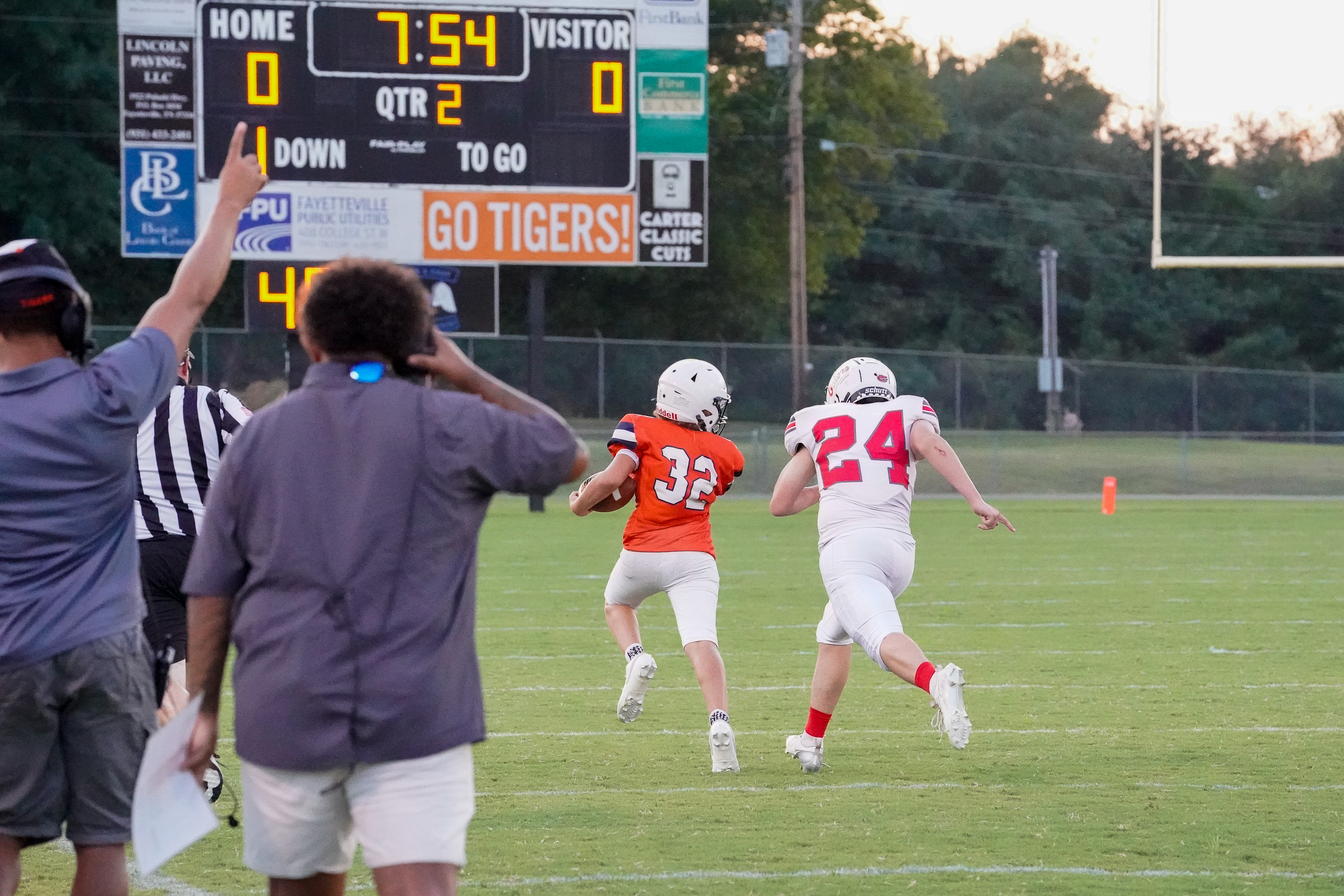 Two young football players, one in orange and one in white, run across a grassy field in front of a scoreboard displaying the game's progress, with trees and a goalpost visible in the background.