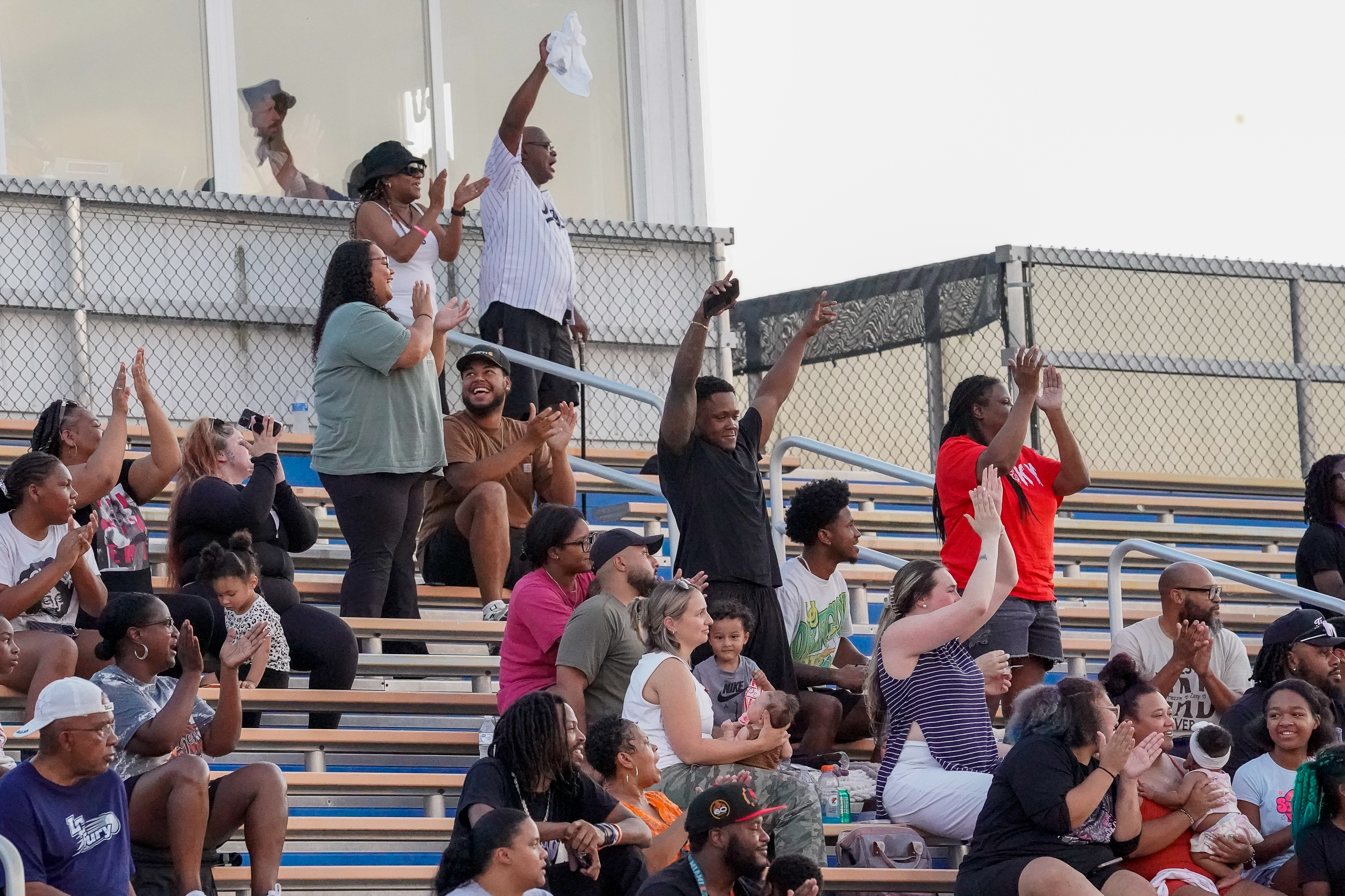 A crowd of people in the foreground cheers from bleachers with a fence and building in the background.