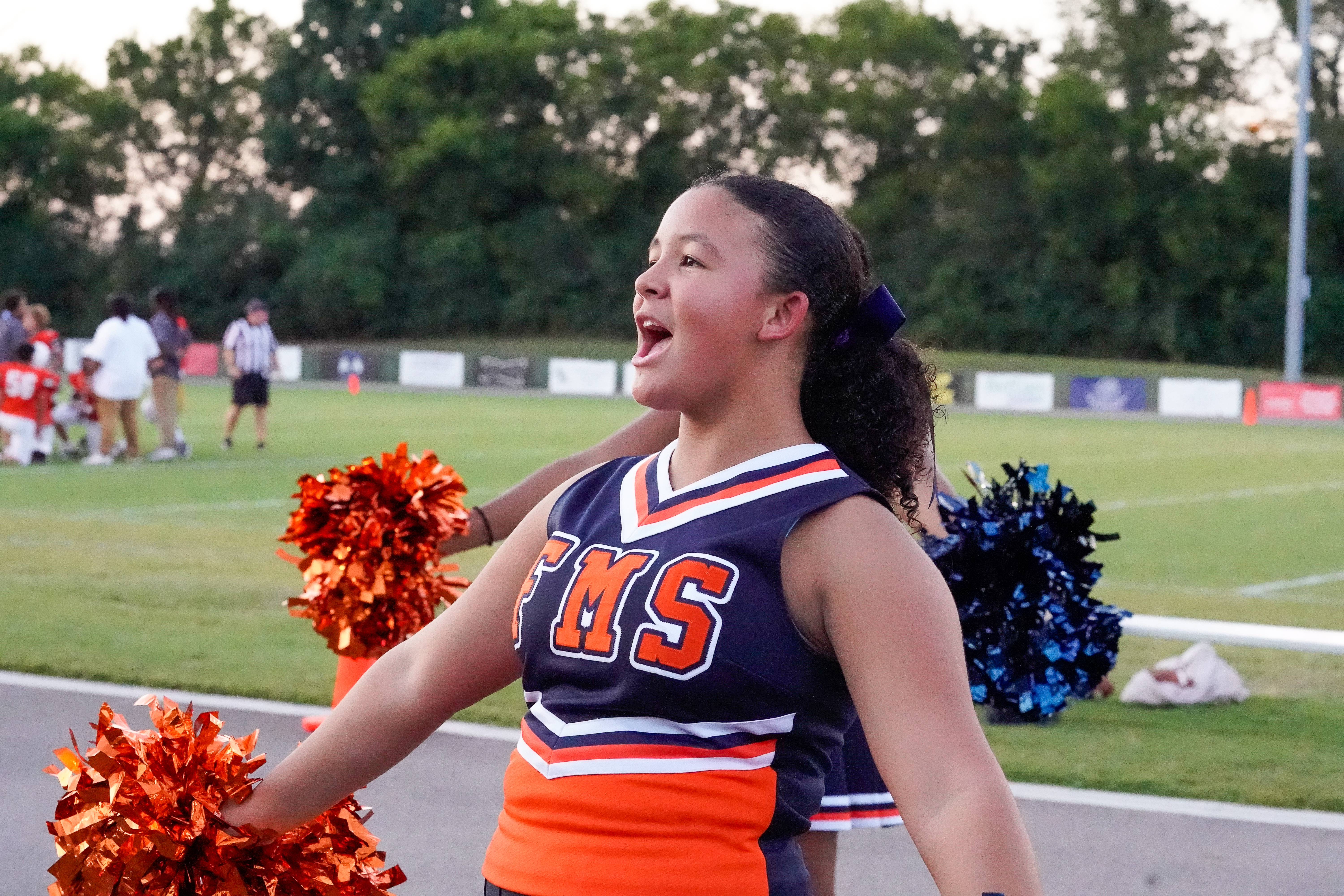 A young cheerleader with orange pom-poms in the foreground cheers on a football field in the background.