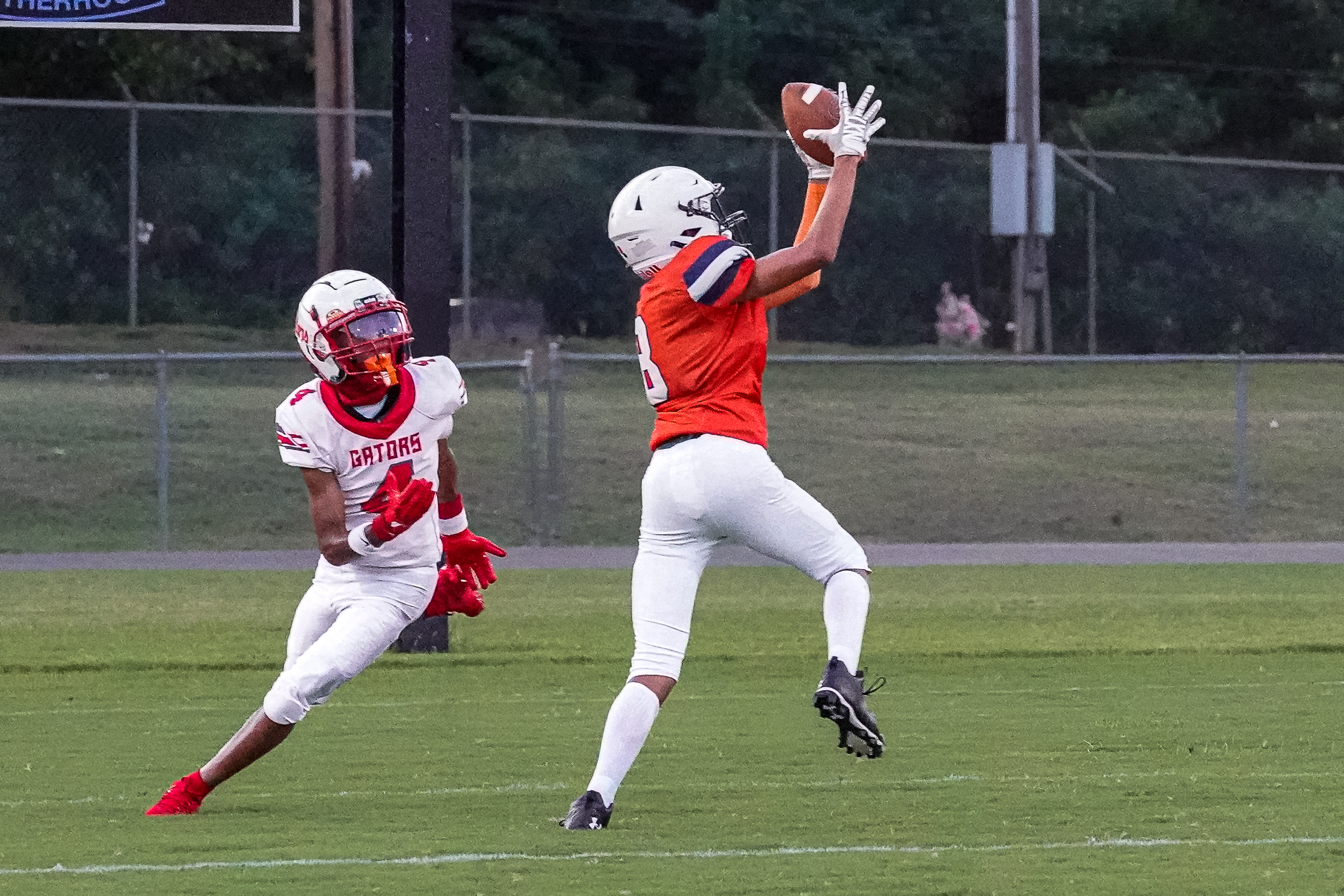 Two football players, one in white and red, the other in orange and white, are on a green field with trees and a fence in the background.