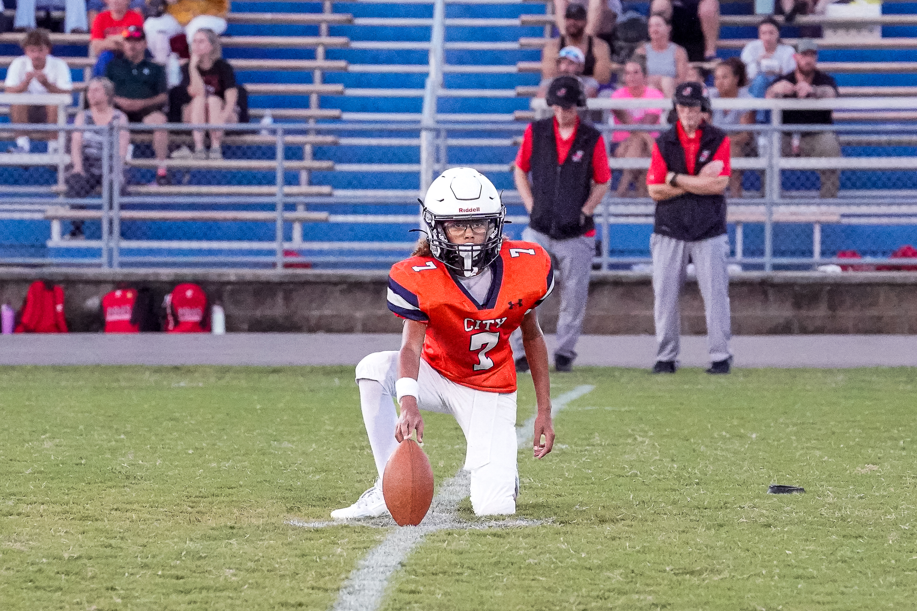 A football player in an orange jersey kneels on a grassy field with a football at his feet, while spectators watch from bleachers in the background.