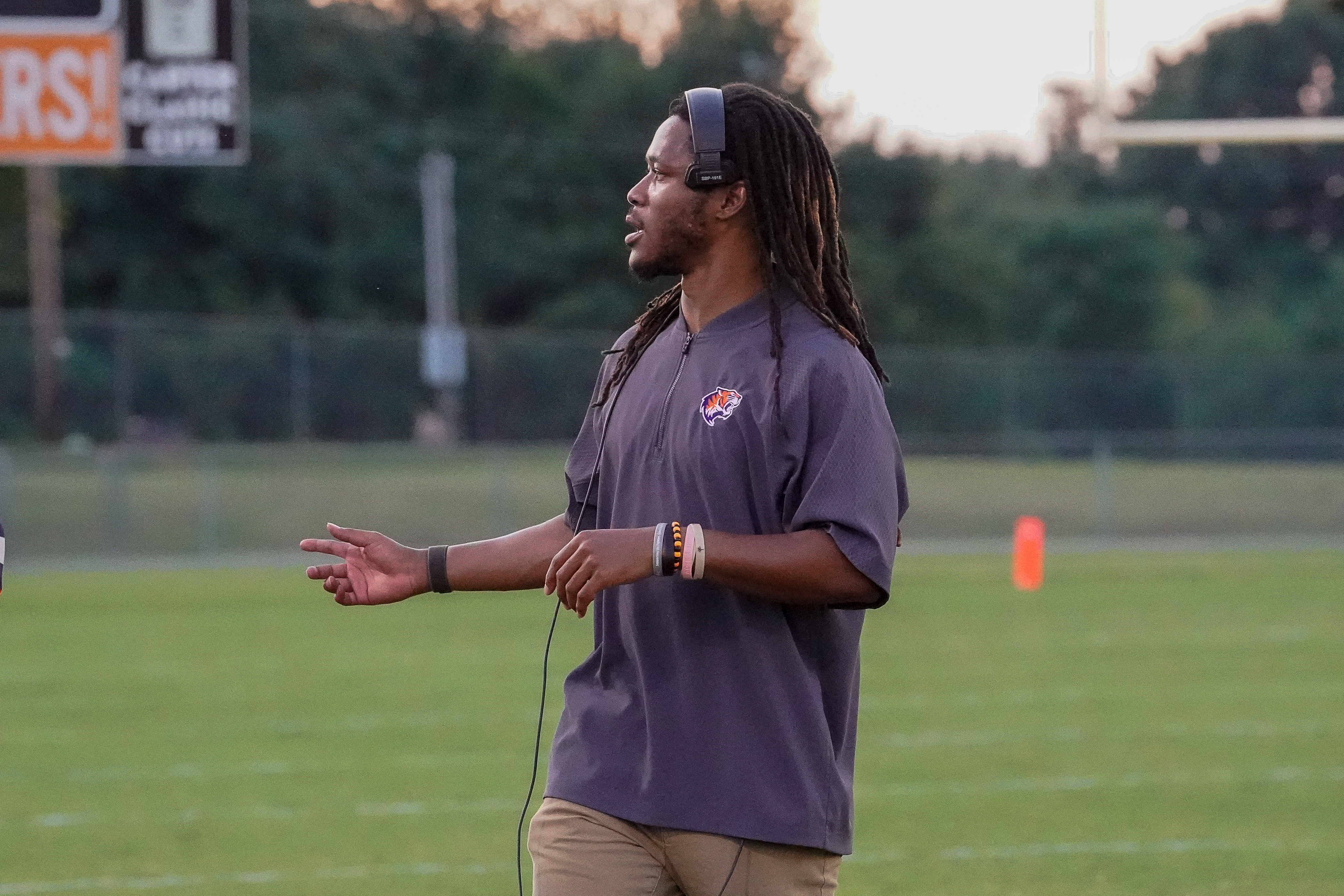 A man with dreadlocks wearing a headset and a gray jacket stands on a grassy football field, with a scoreboard and trees visible in the background.