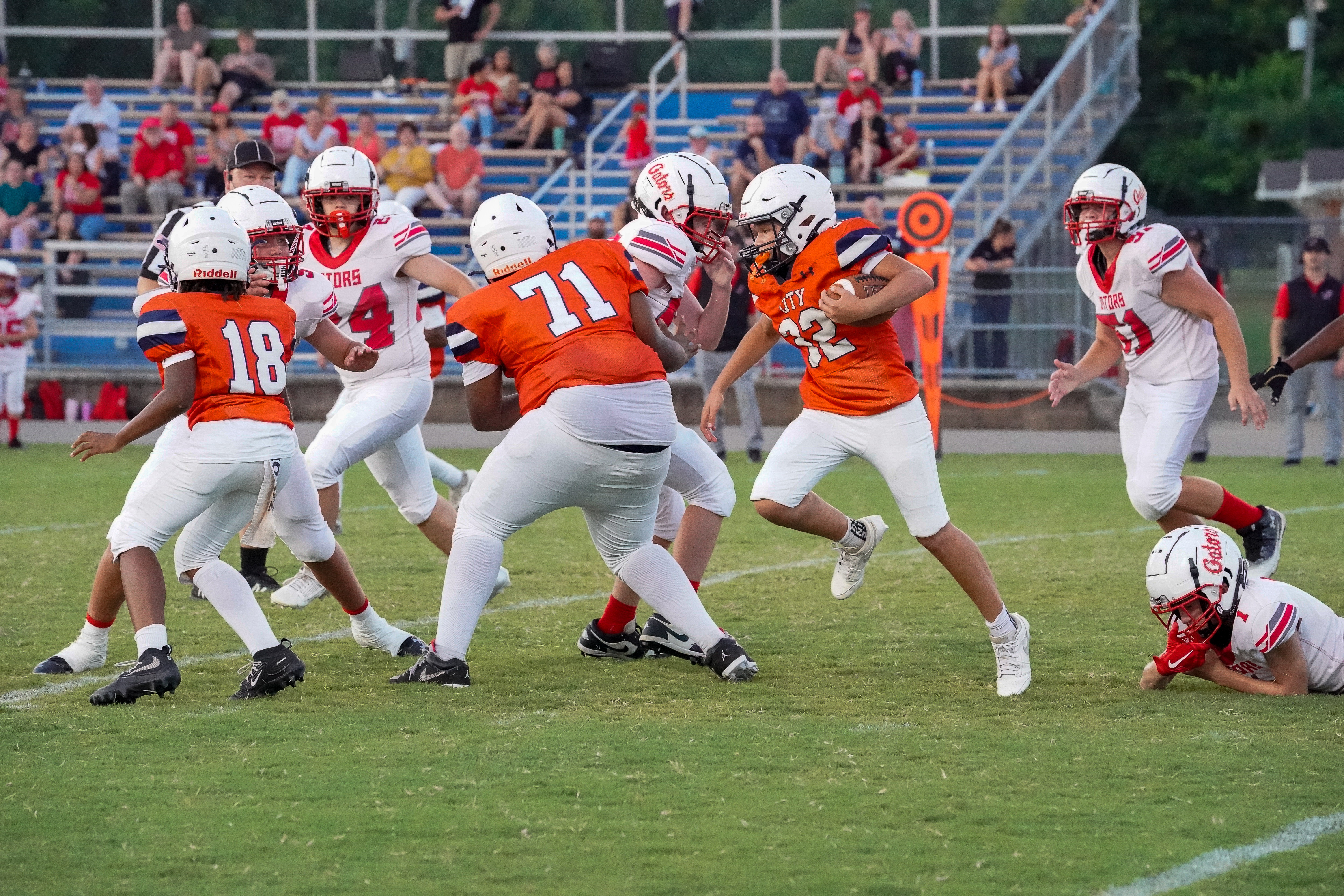 Football players in orange and white uniforms are engaged in a play on a grassy field, with spectators in blue bleachers in the background.