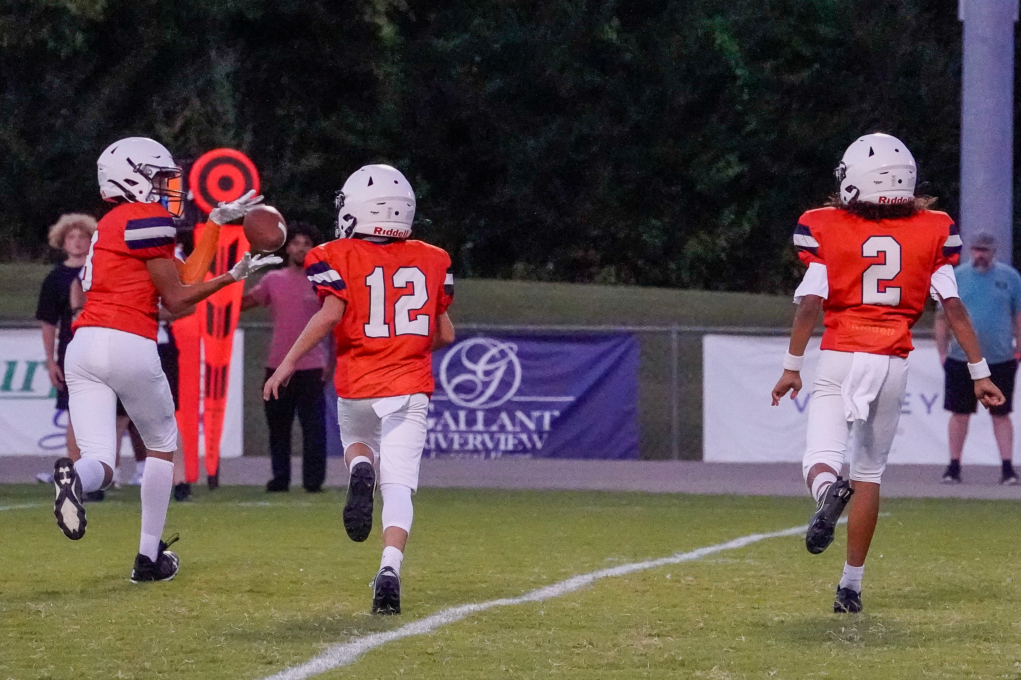 Three young football players in orange jerseys run across a grassy field with a stadium and trees in the background.