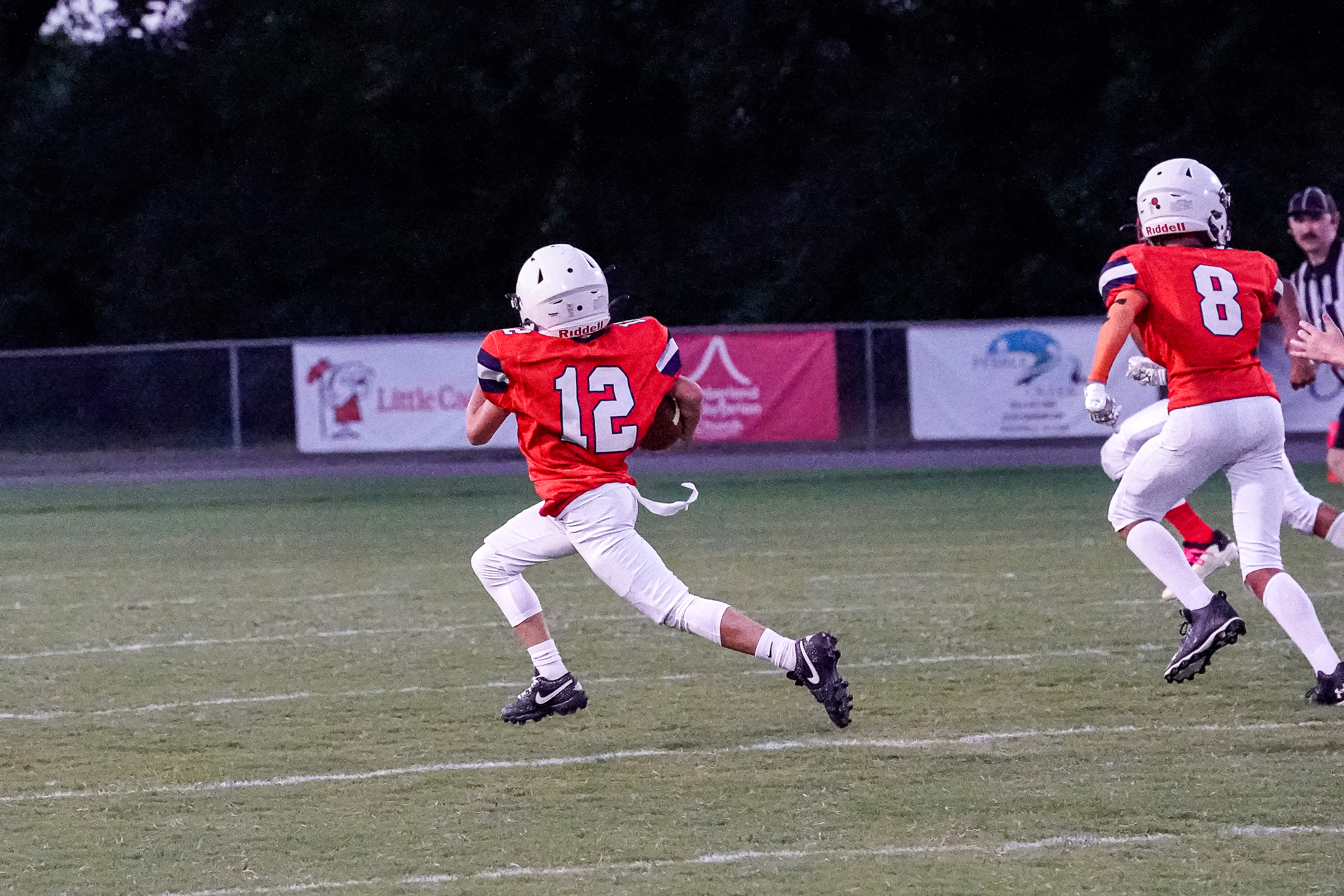 A young football player in an orange jersey with the number 12 runs with the ball across a grassy field, while other players and a fence with advertisements are visible in the background.