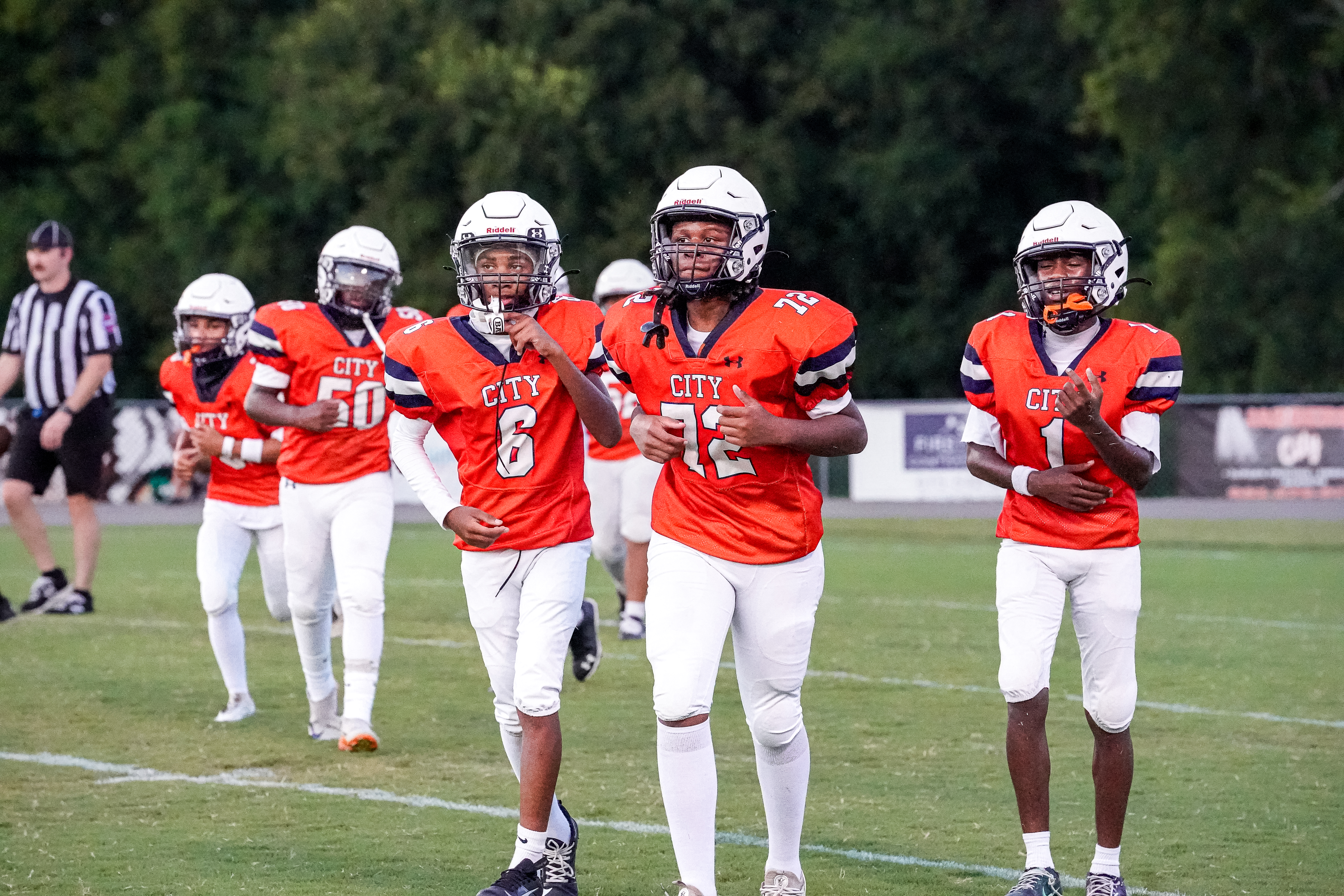 A group of young football players in orange jerseys run across a green field with trees and a fence in the background.