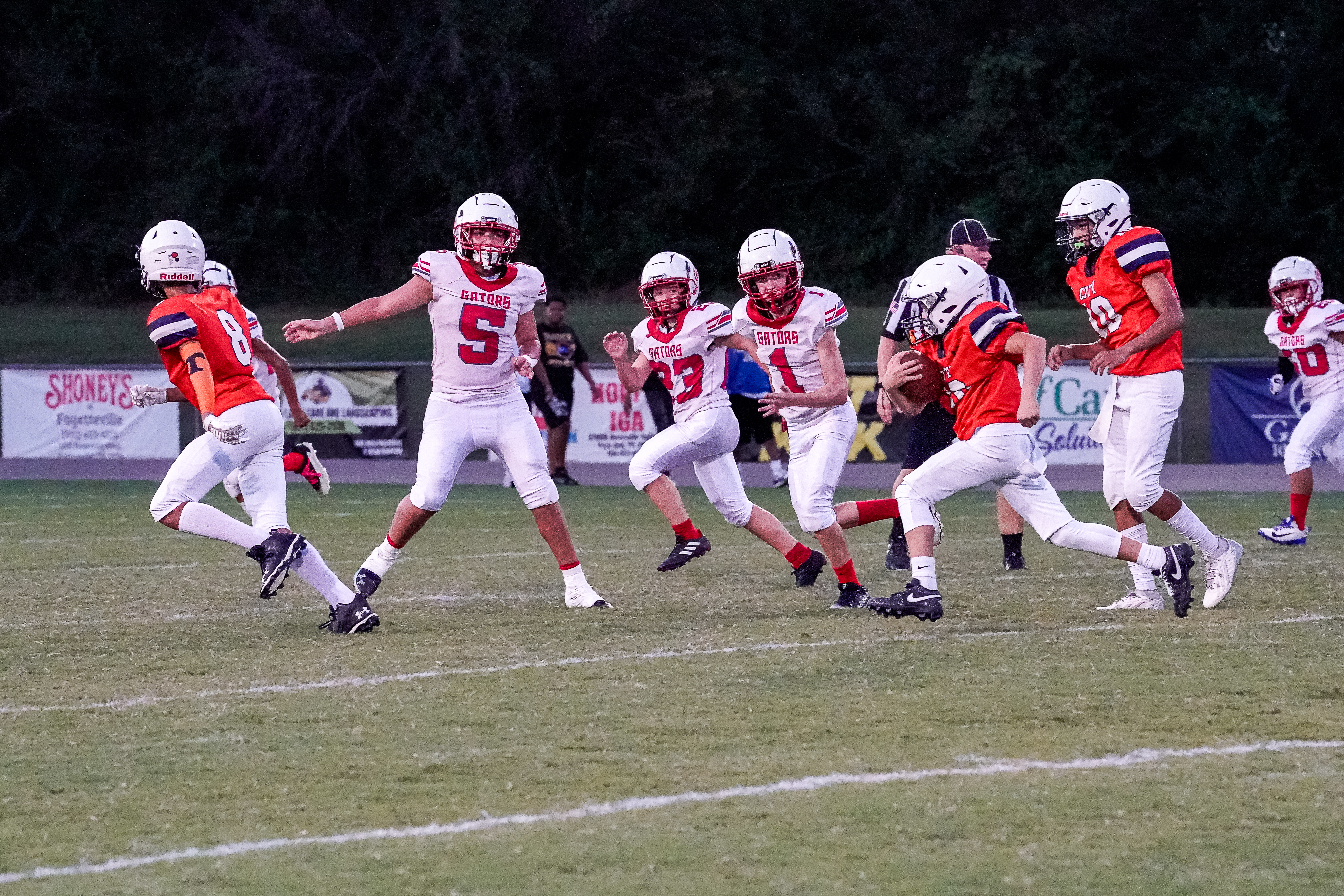Football players in red and white uniforms run across a grassy field with trees and stadium lights in the background.