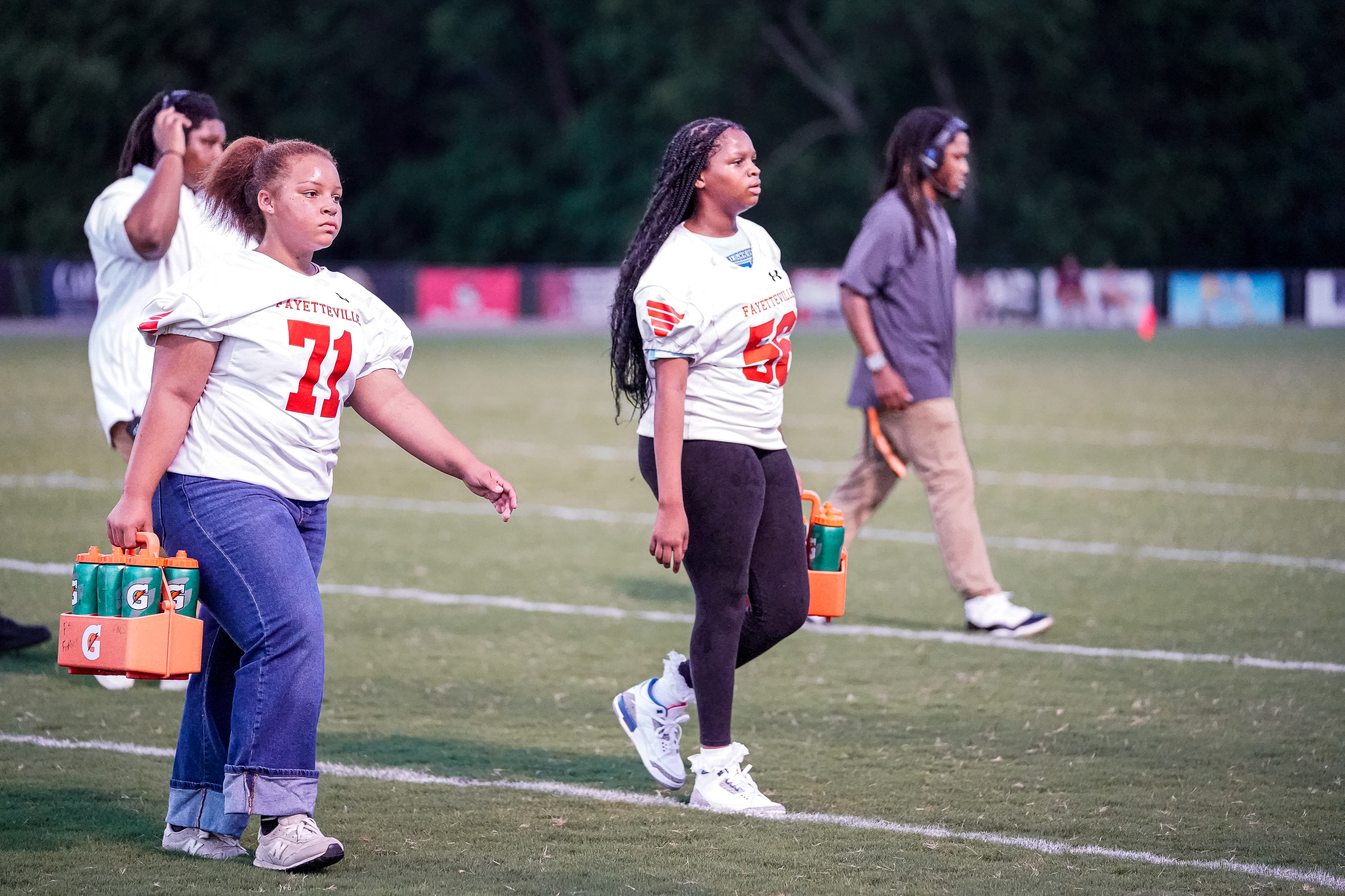 Two women in football jerseys walk across a grassy field carrying drinks, with a blurred background of trees and stadium lights.
