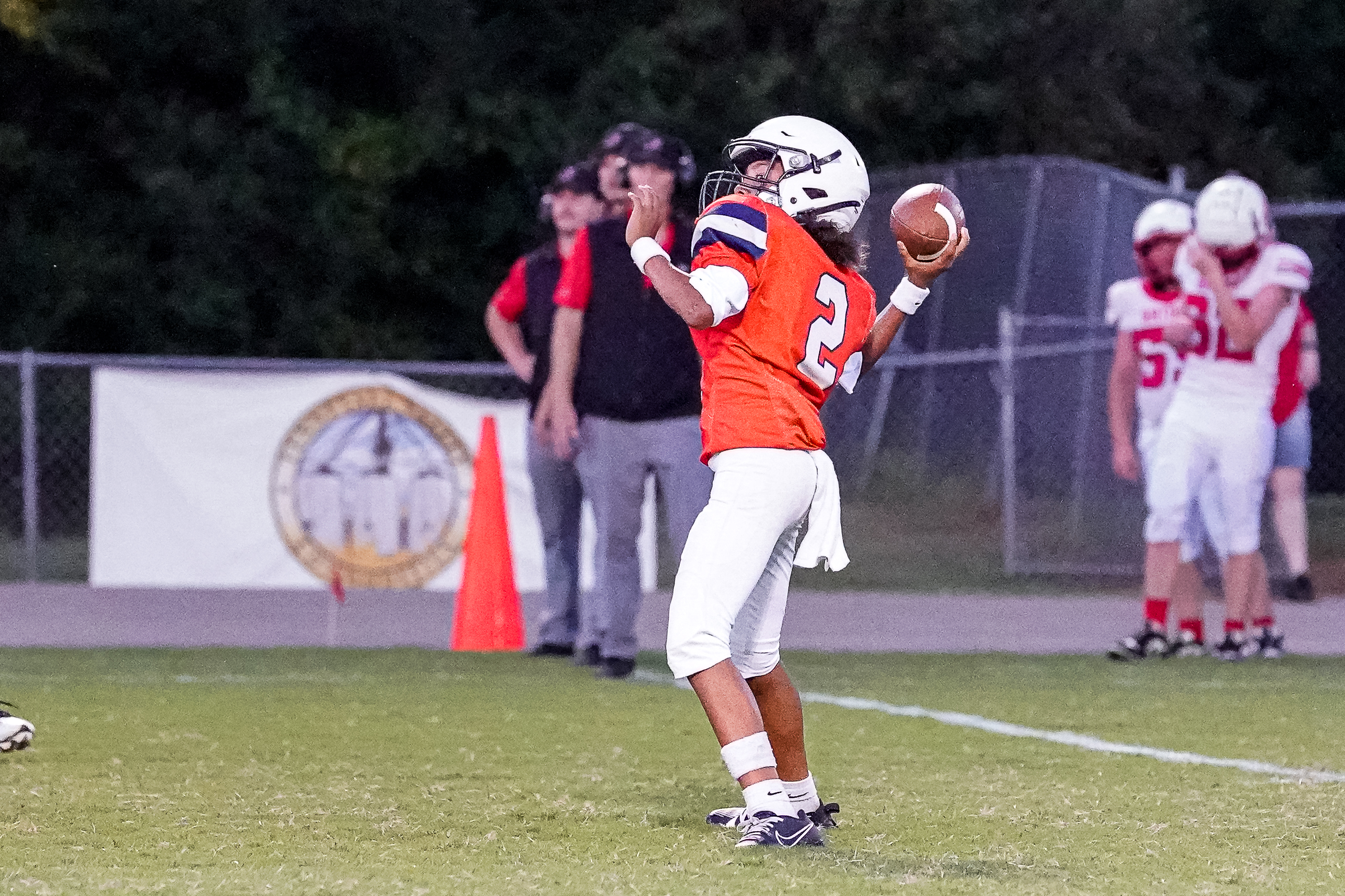 A football player in an orange jersey throws a pass on a field with spectators and a banner in the background.