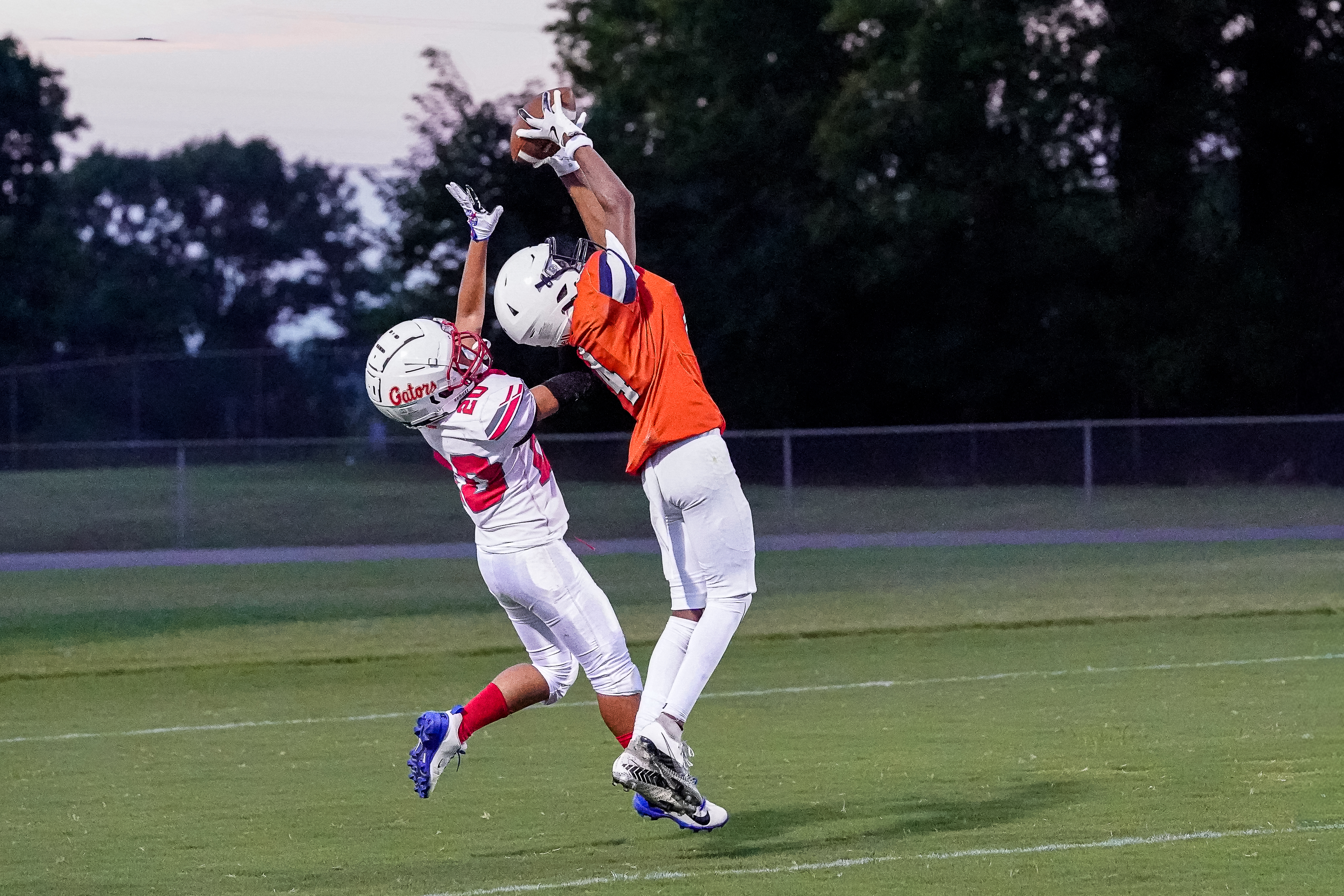 Two football players in mid-air leap for a pass against a backdrop of trees and a dusky sky.