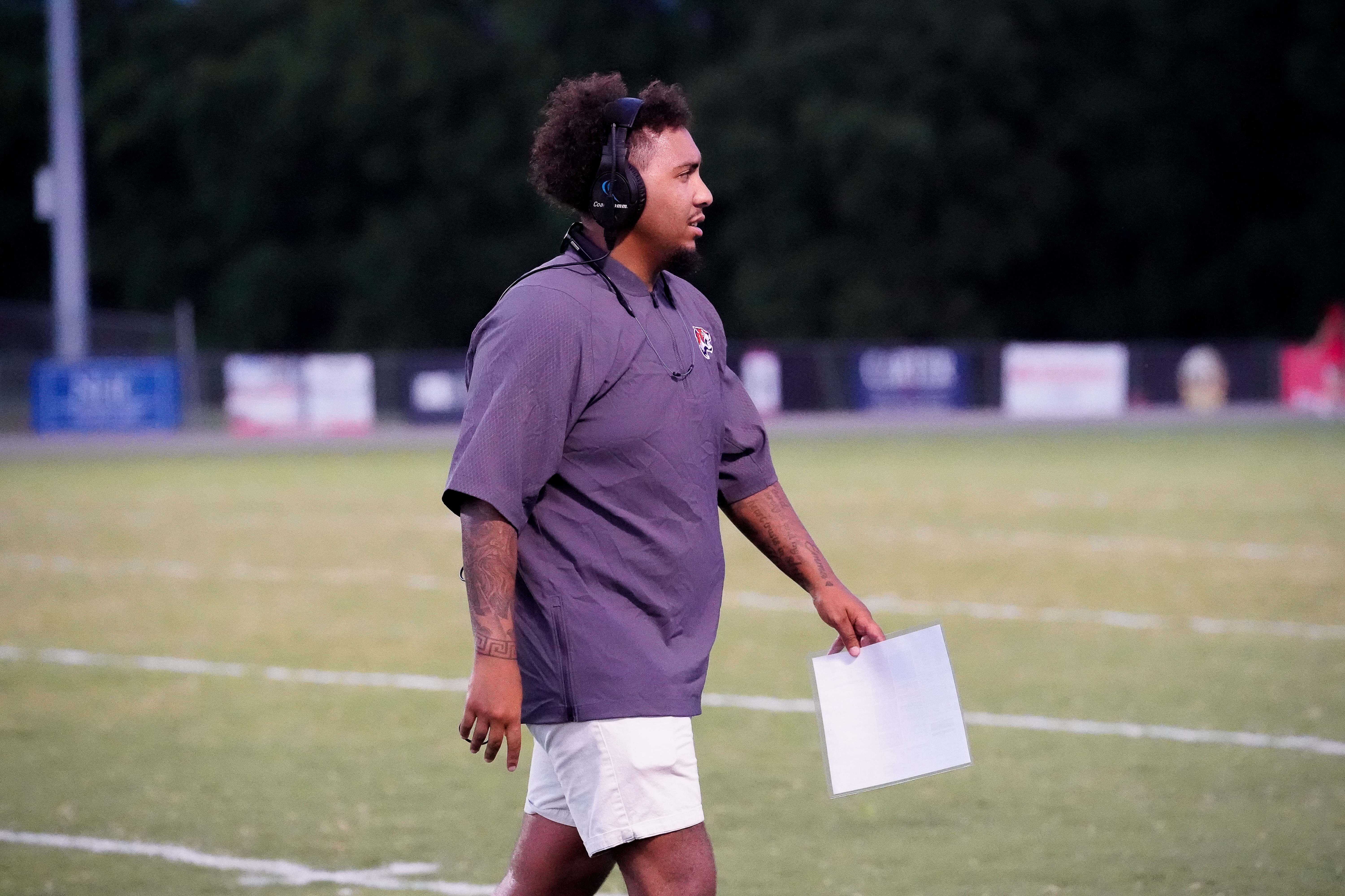A man in a headset and shorts walks across a football field, with stadium lights and trees in the background.