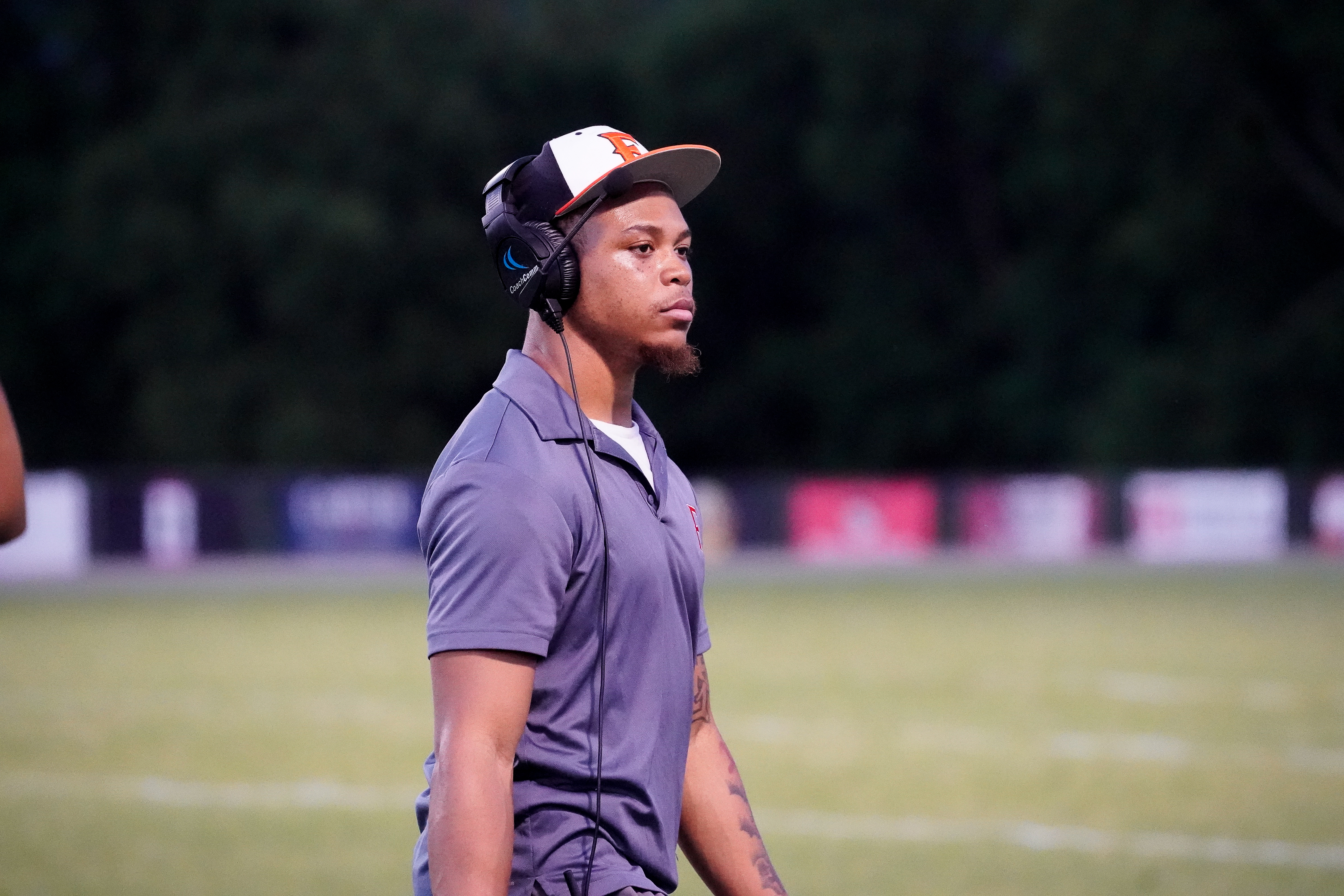 A young man wearing a headset and a cap stands on a grassy field with trees in the blurred background.