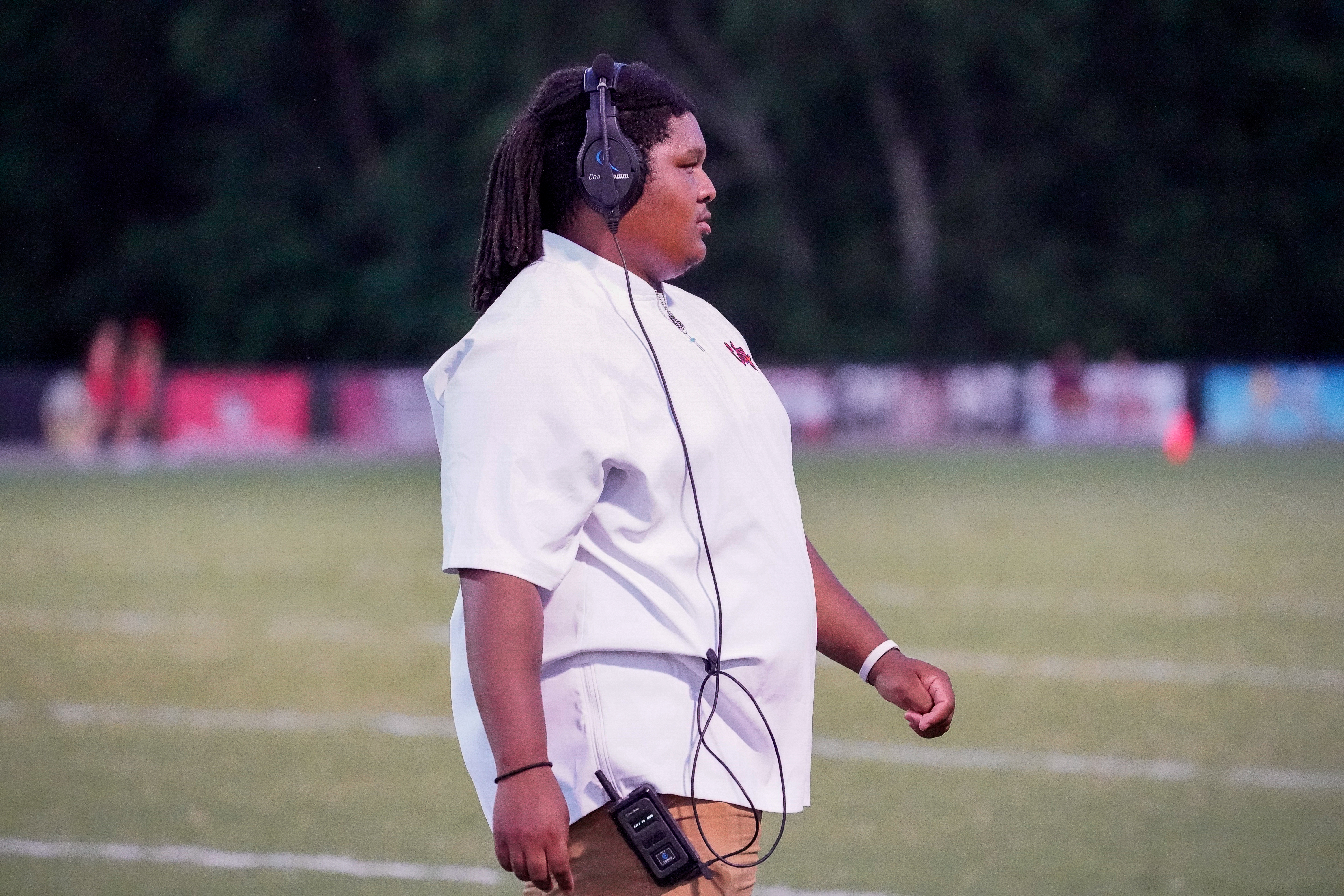 A young person wearing headphones and a white shirt stands on a grassy field with a blurred background of a football game.