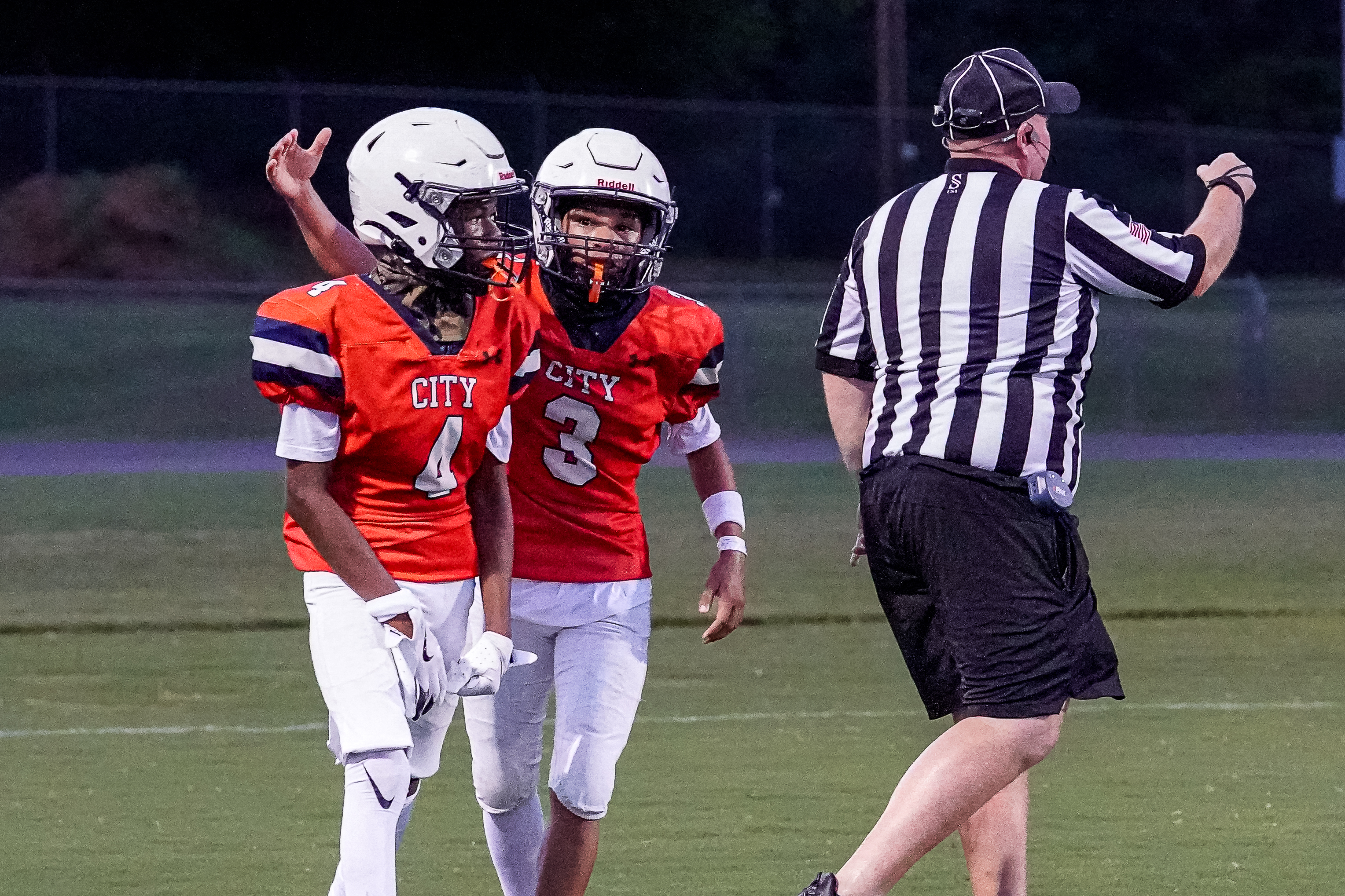 Two young football players in orange jerseys celebrate on a green field while a referee in a striped shirt stands to the right.