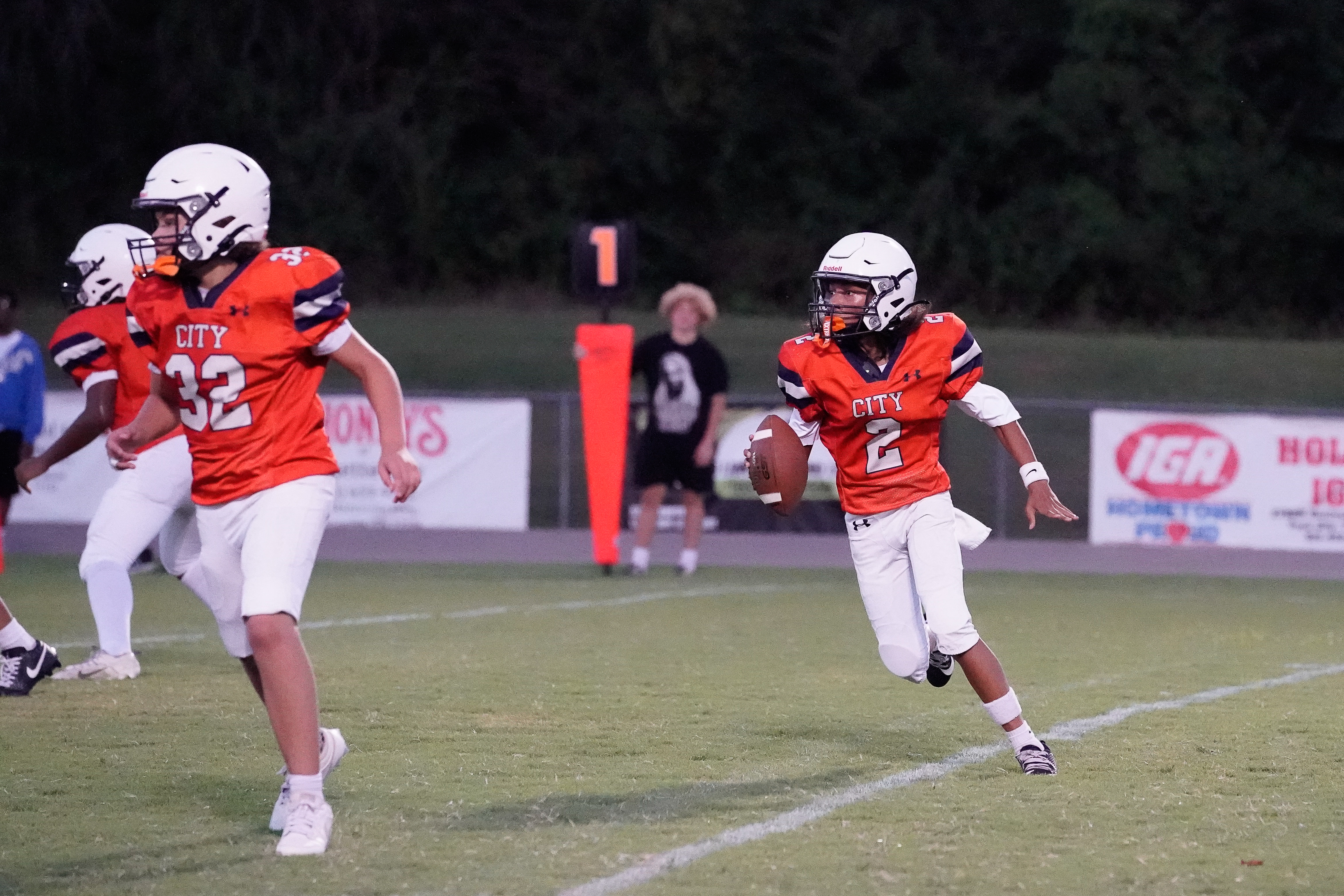 Two young football players in orange jerseys run on a grassy field with a scoreboard and spectators in the background.
