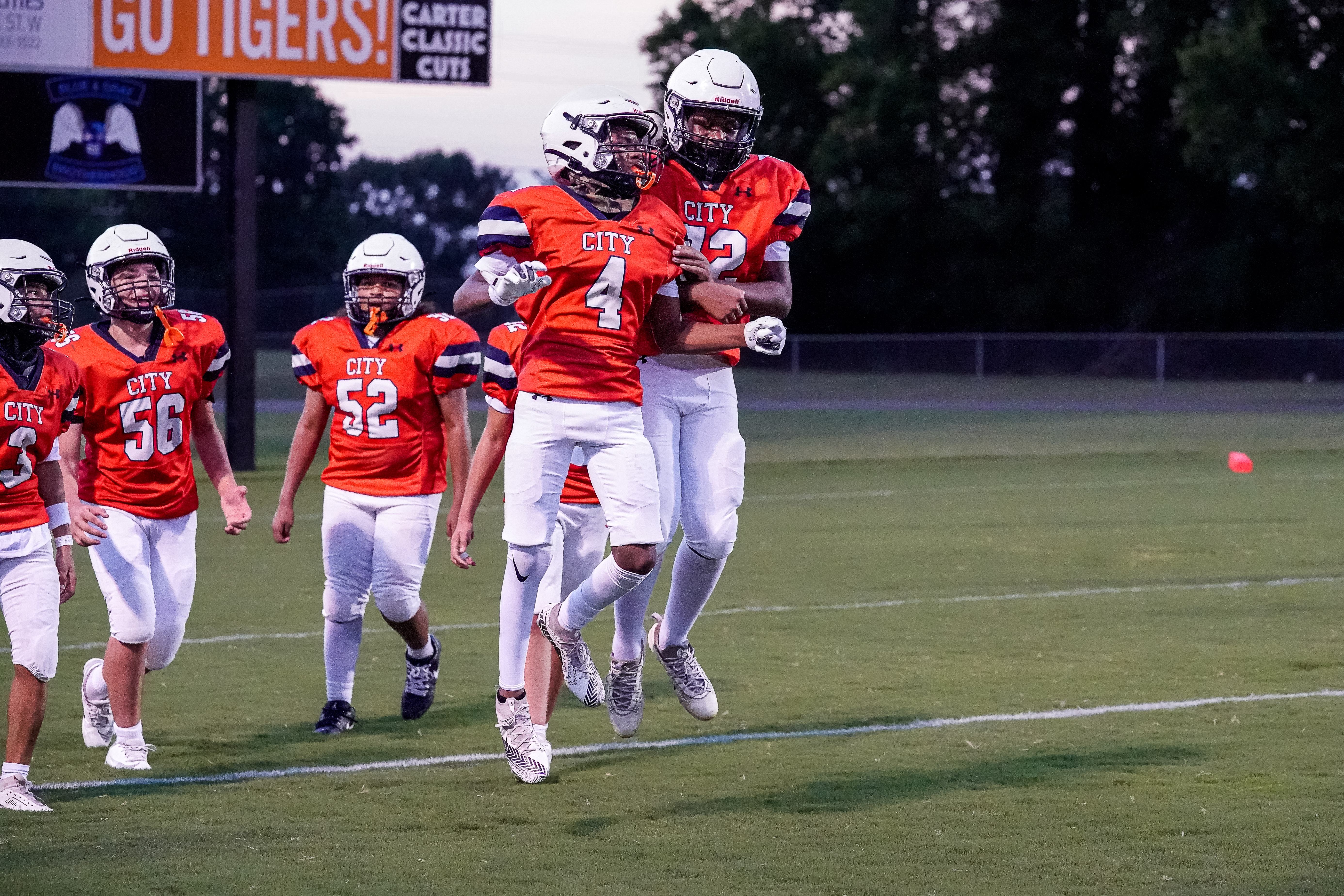 A group of young football players in orange jerseys celebrate on a grassy field with a scoreboard in the background.