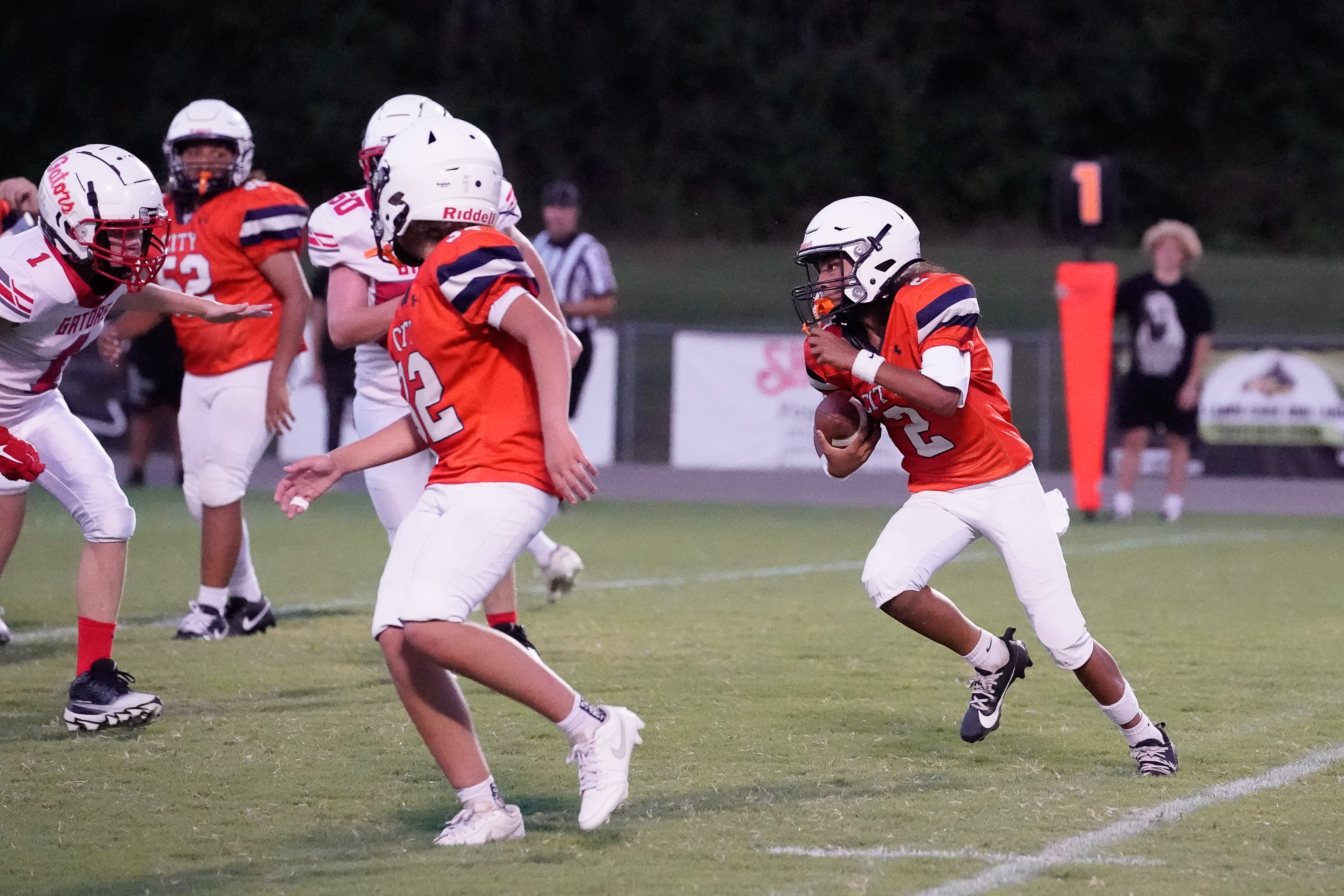 In the foreground, young football players in orange and white uniforms run on a grassy field, while in the background, spectators watch the game under the dim evening light.
