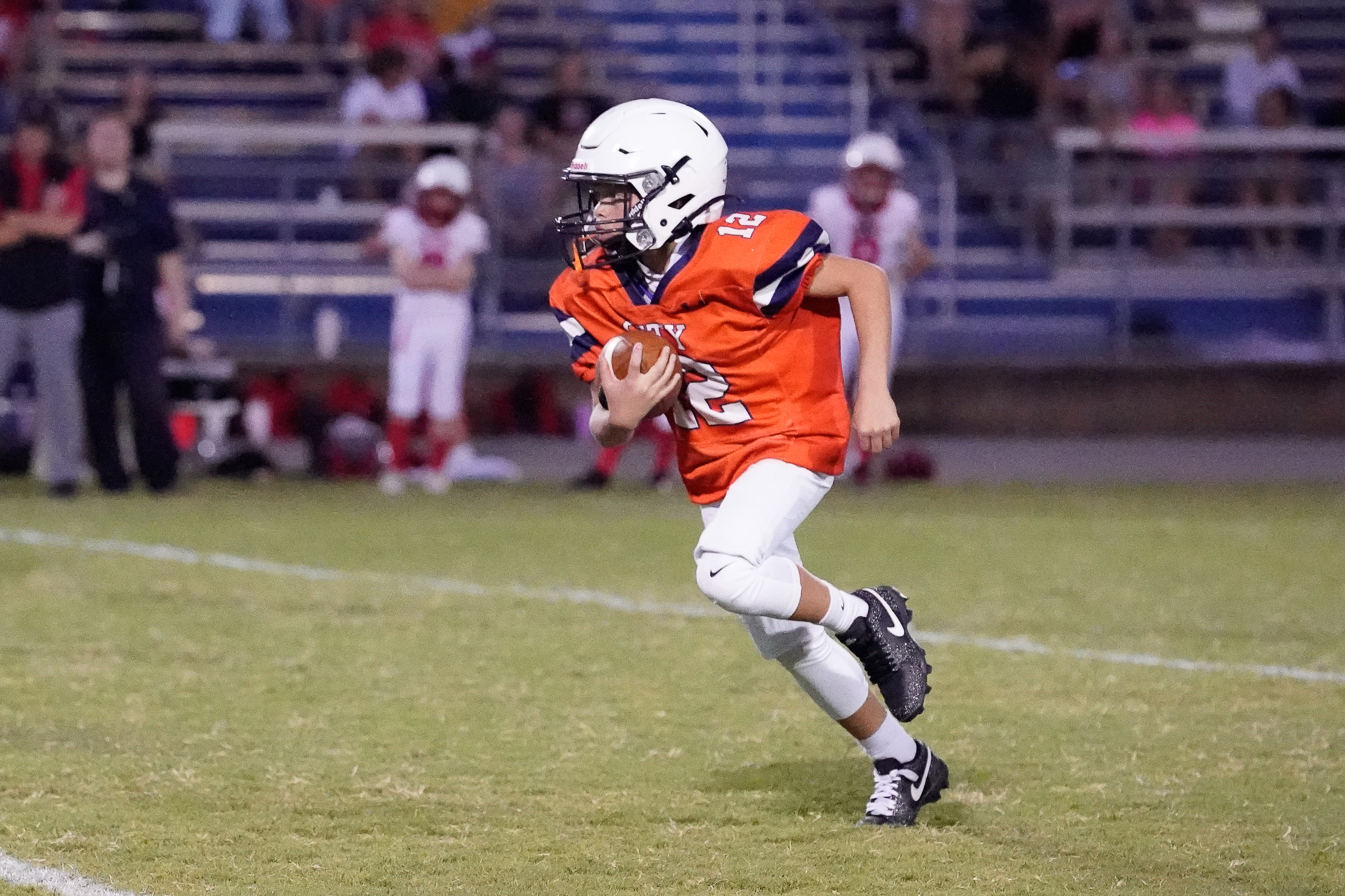 A young football player in an orange jersey runs with the ball on a grassy field, with spectators in the bleachers behind him.