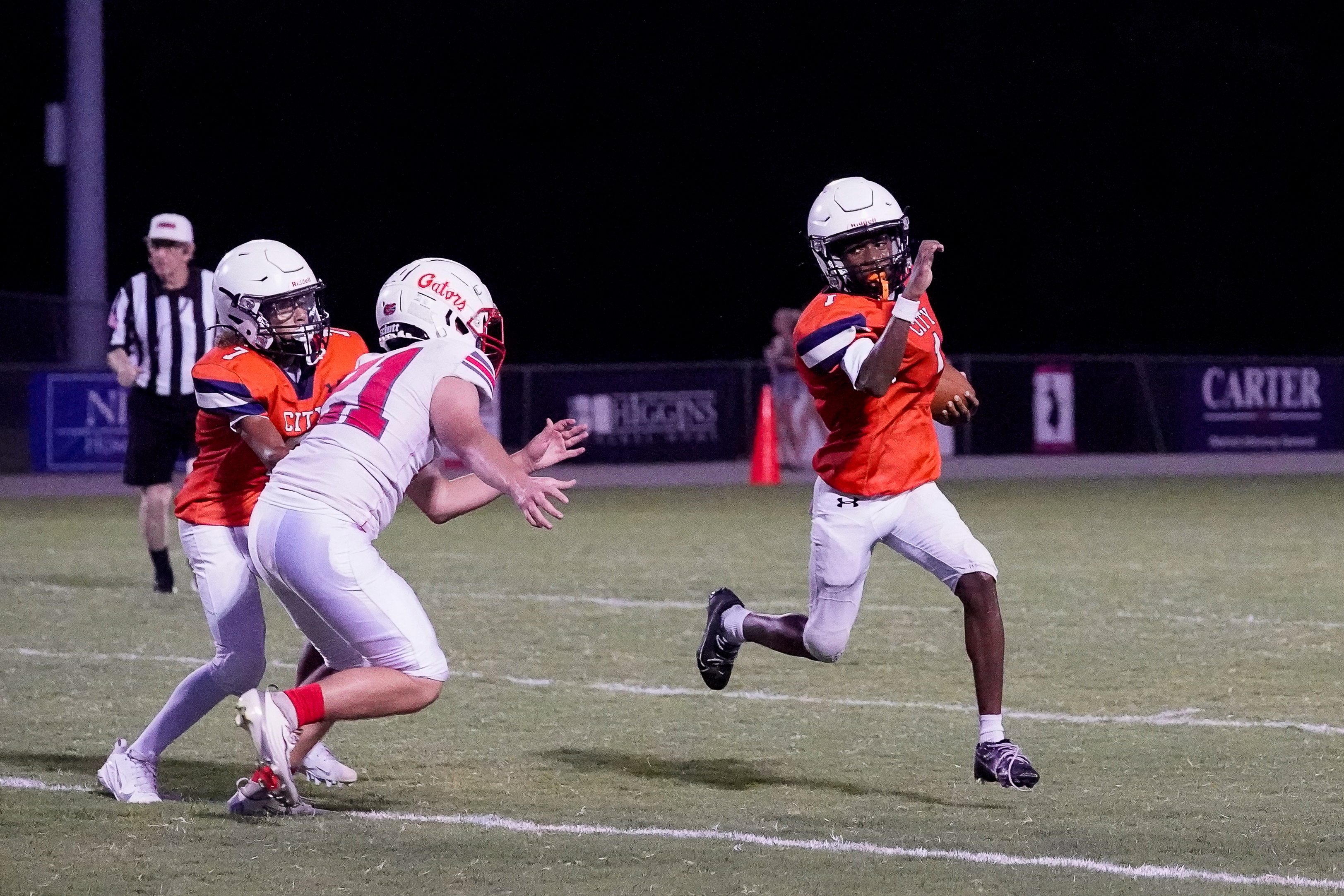 A football player in an orange jersey runs with the ball while two defenders in white jerseys attempt to tackle him, with a dark night sky and stadium lights in the background.
