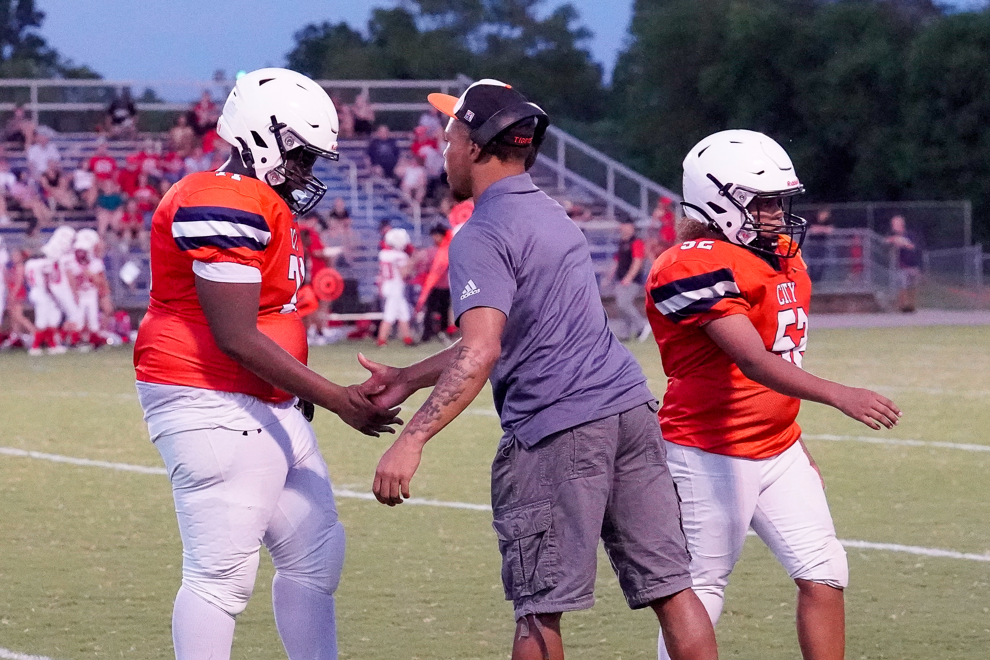 Two football players in orange jerseys and white helmets shake hands with a coach in a grey shirt and cargo shorts on a field, with spectators in the stands behind them.