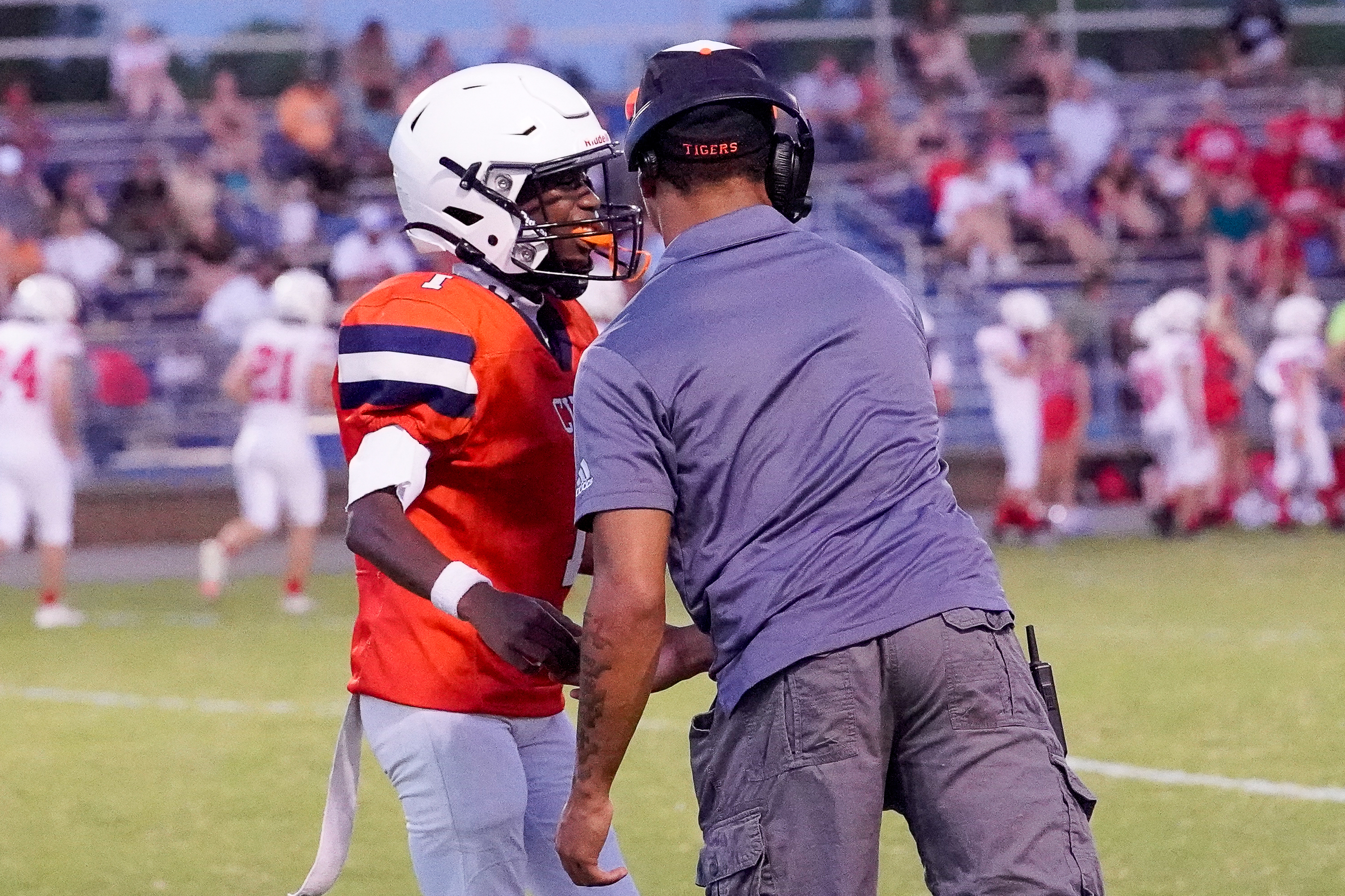A football player in an orange jersey and white helmet is being spoken to by a coach on a field, with spectators in the stands behind them.