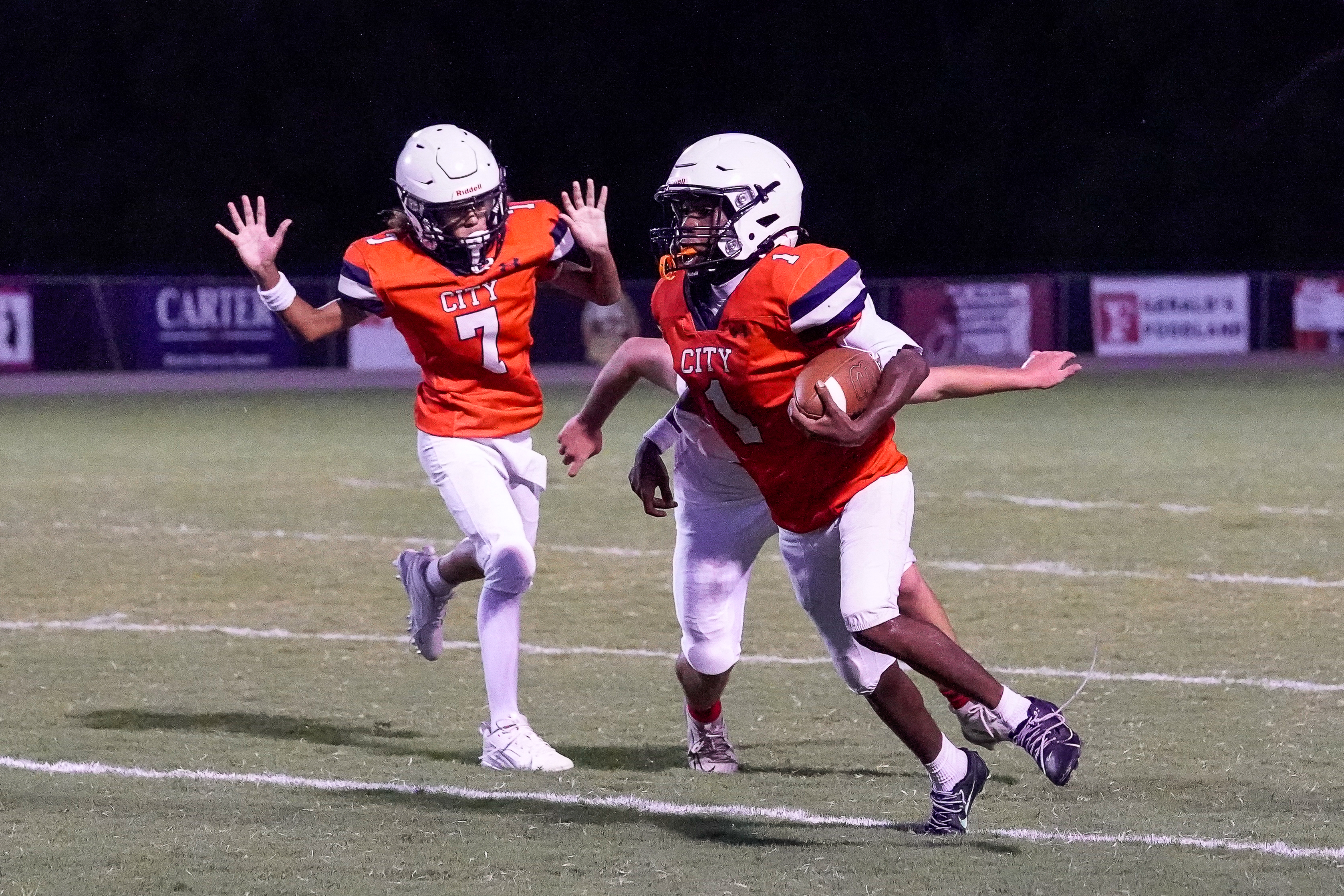 Two football players in orange jerseys run on a field at night, with stadium lights and advertisements visible in the dark background.