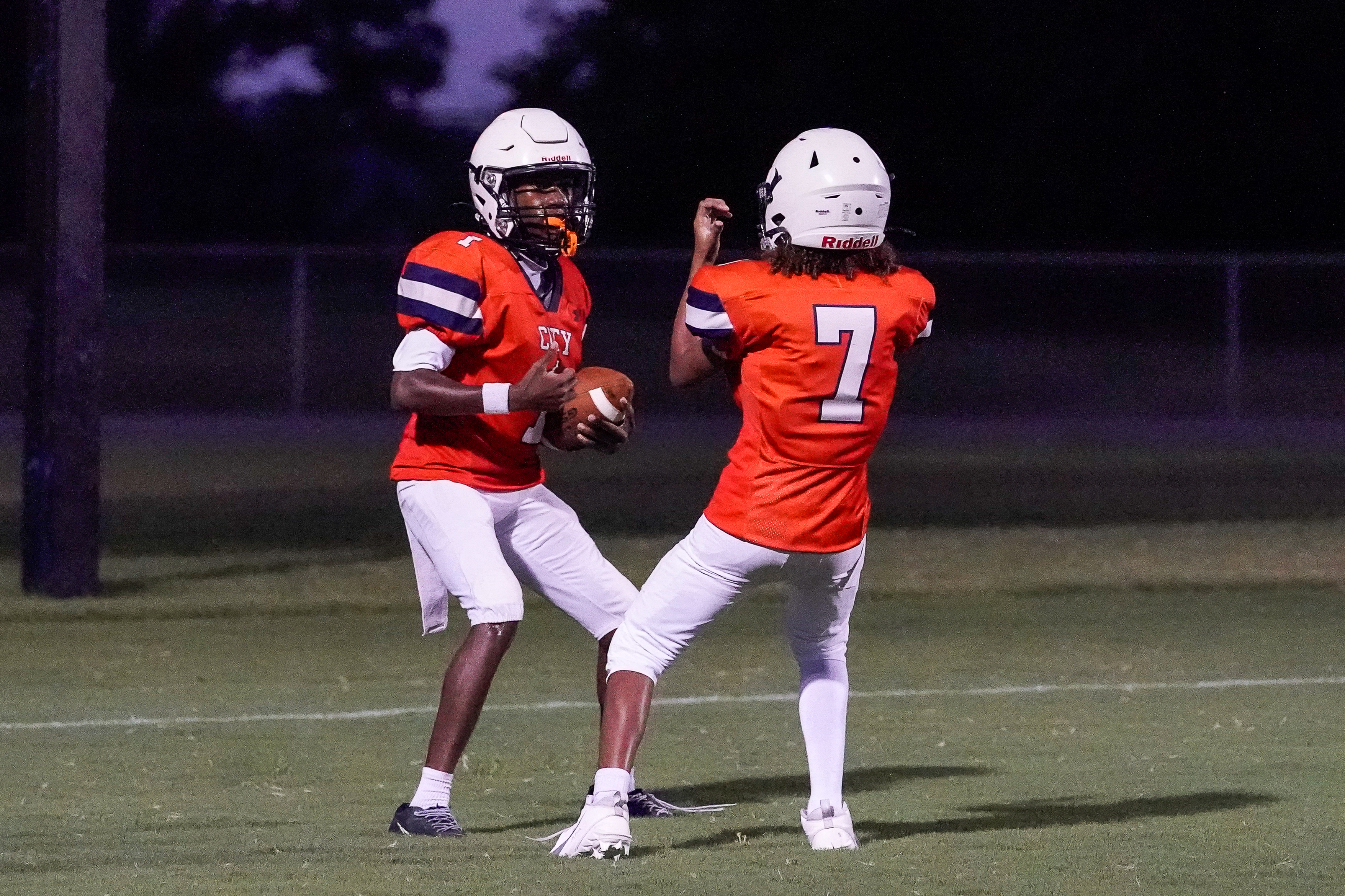 Two football players in orange jerseys and white pants stand on a grassy field at dusk, with a fence and trees visible in the blurred background.