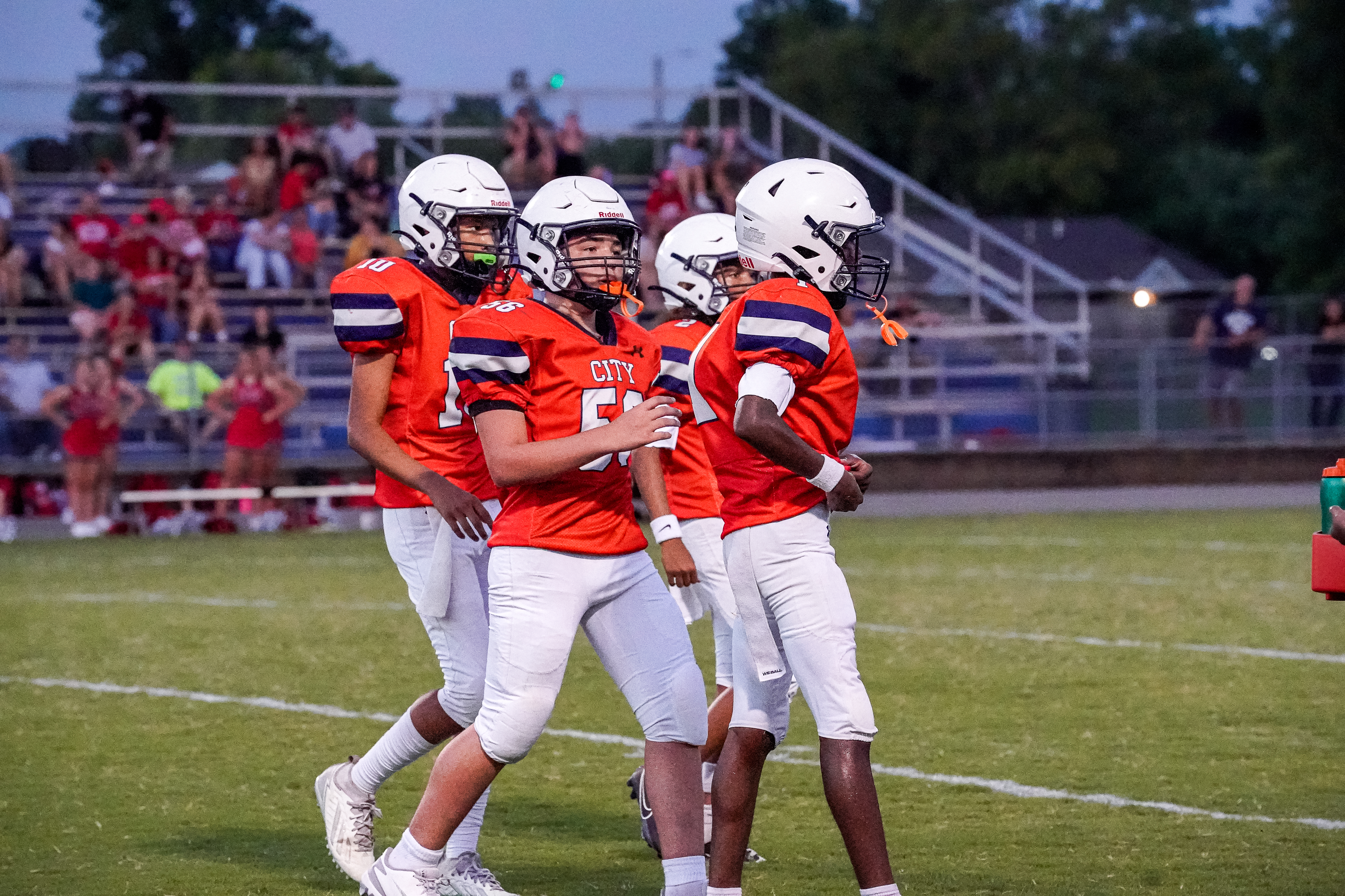 Four young football players in orange jerseys and white helmets stand on a grassy field in the foreground, while a crowd watches from bleachers in the blurred background.