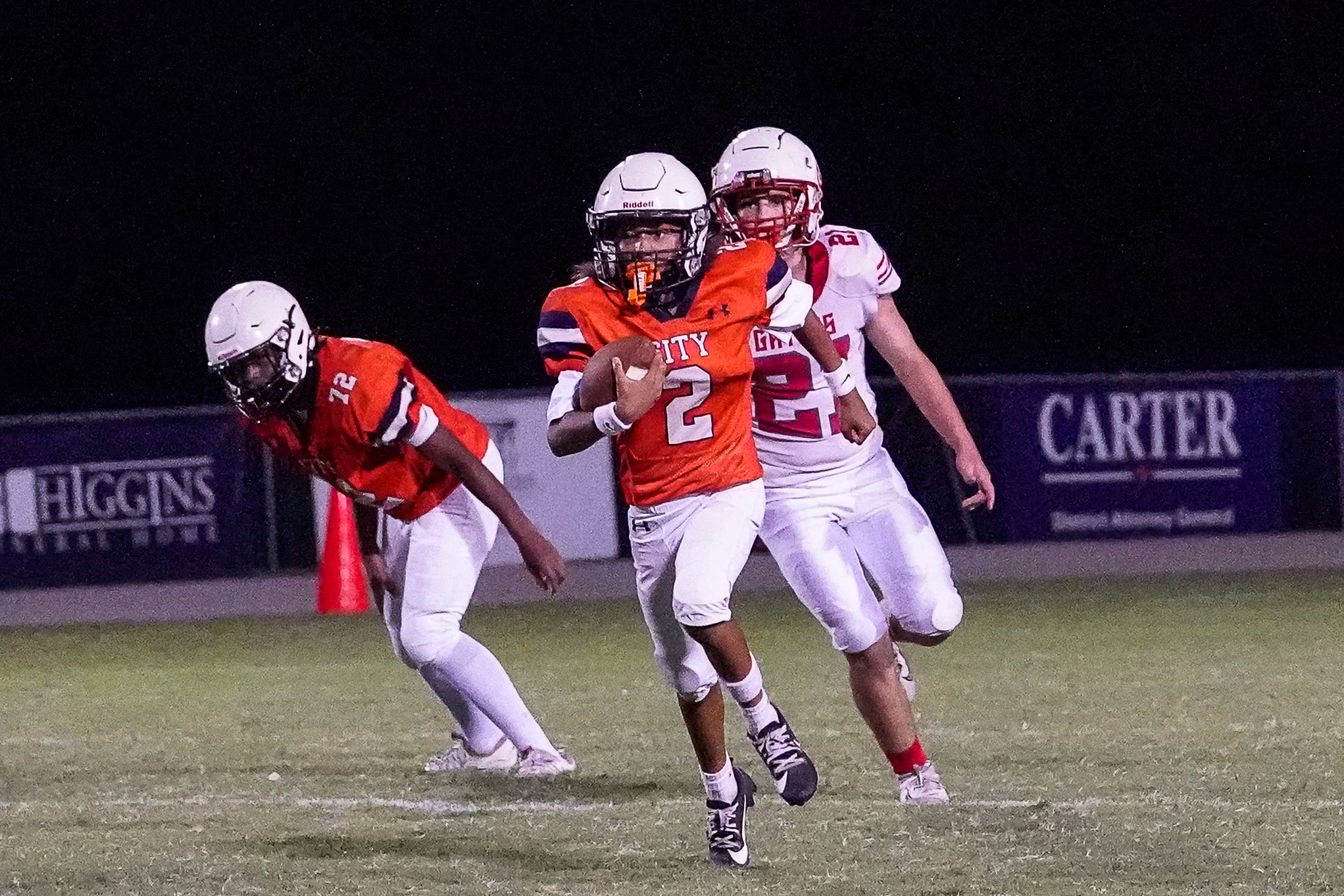 A football player in an orange jersey with the number 2 runs with the ball in the foreground, while other players in white and red uniforms and a dark background with sponsor signs are visible behind him.