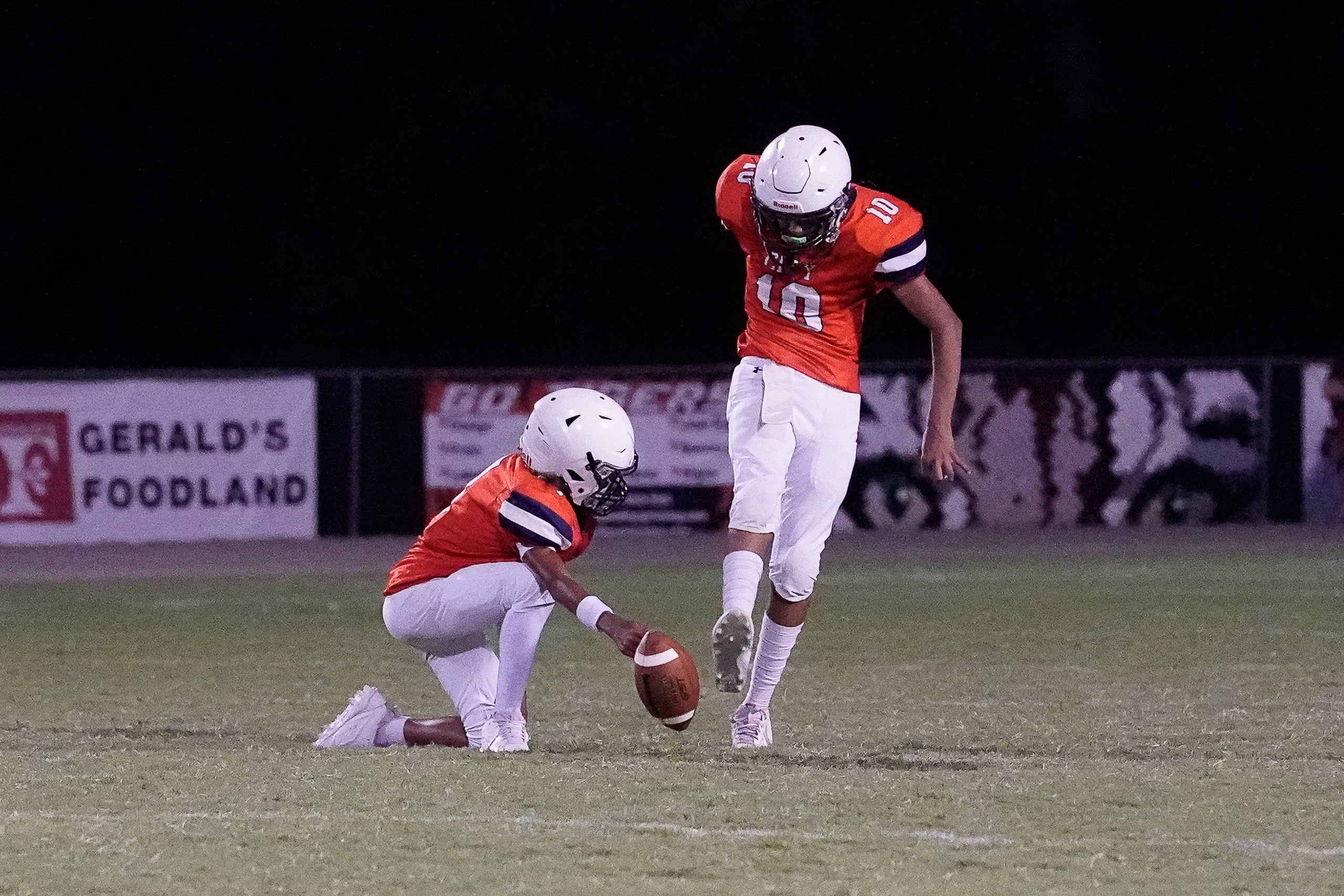 Two football players in orange jerseys are on a field, with one holding the ball for the other to kick, as a banner for "Gerald's Foodland" is visible in the dark background.