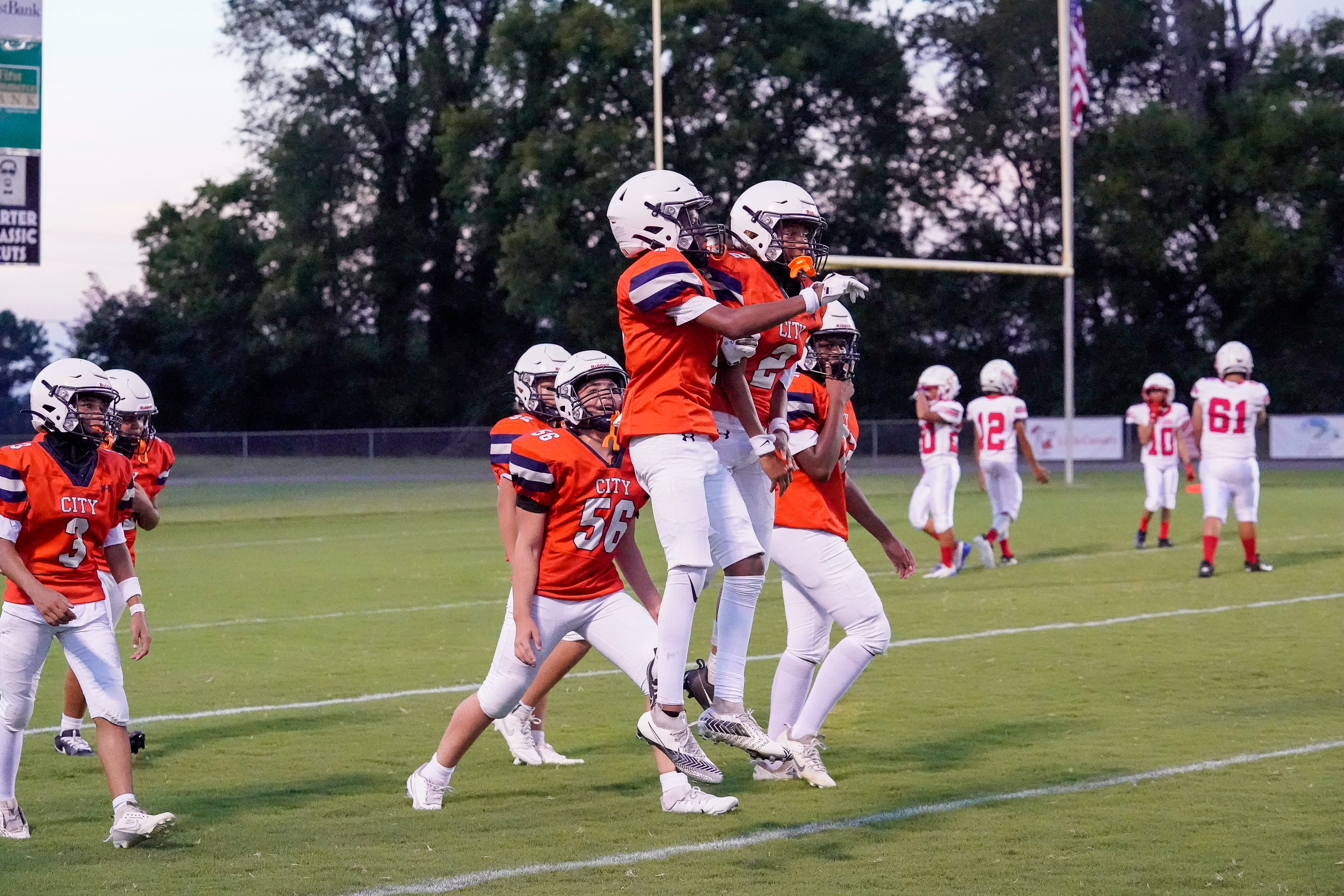 Football players in orange and white uniforms are on a green field with goalposts and trees in the background.