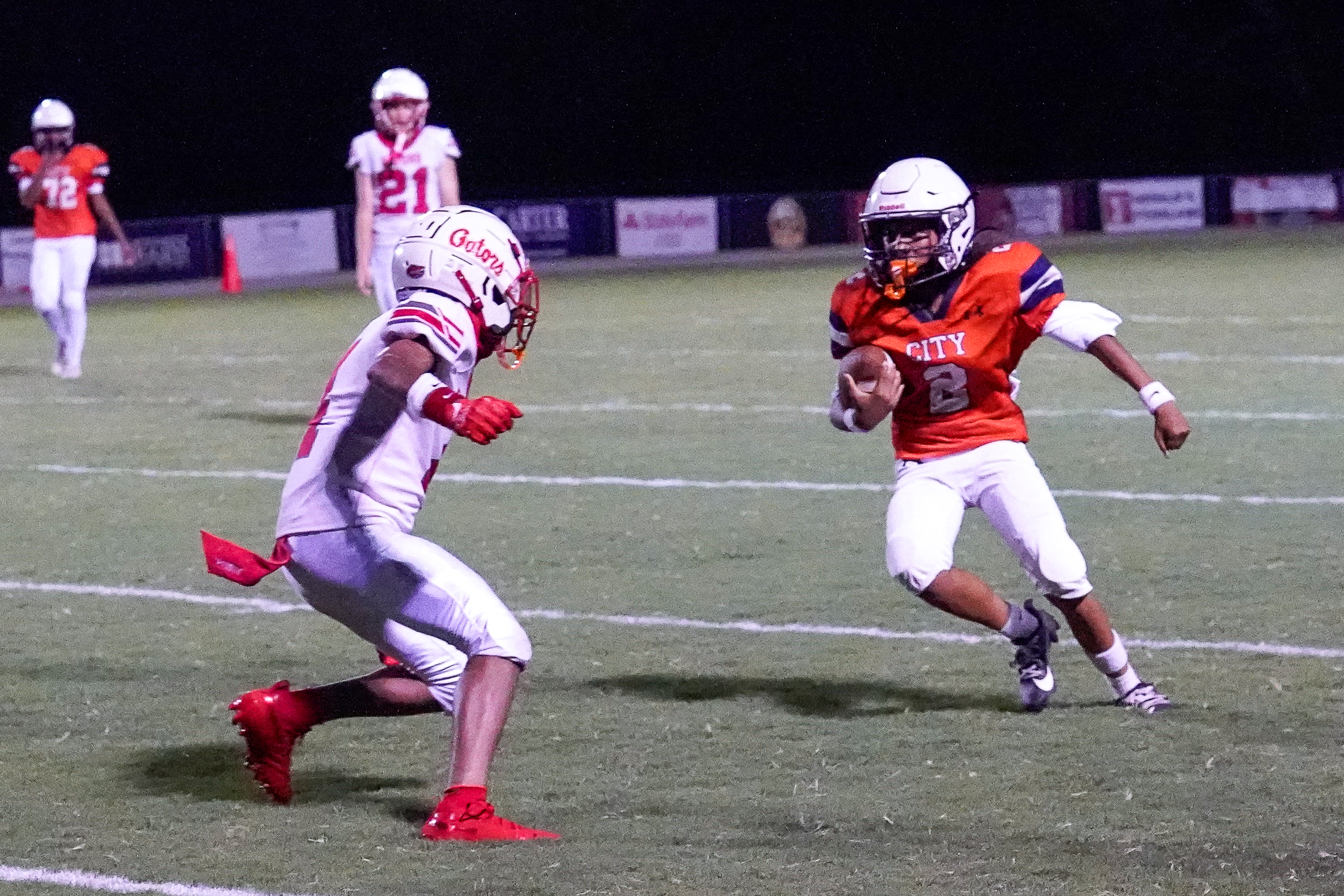 A young football player in an orange jersey runs with the ball while a player in a white jersey attempts to tackle him on a grassy field with spectators in the background.