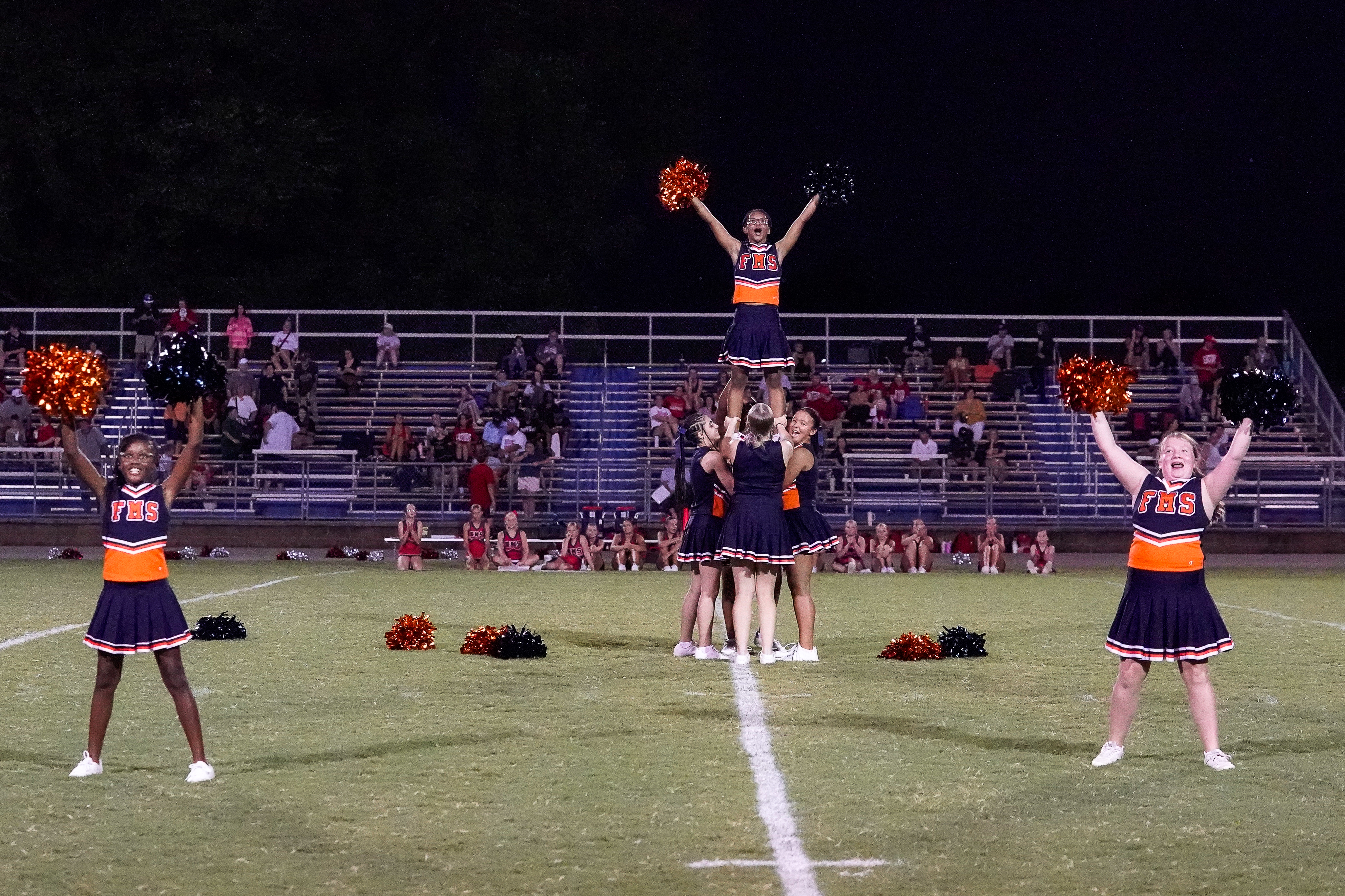 Cheerleaders in orange and navy uniforms perform a pyramid on a grassy football field with spectators in bleachers in the background.