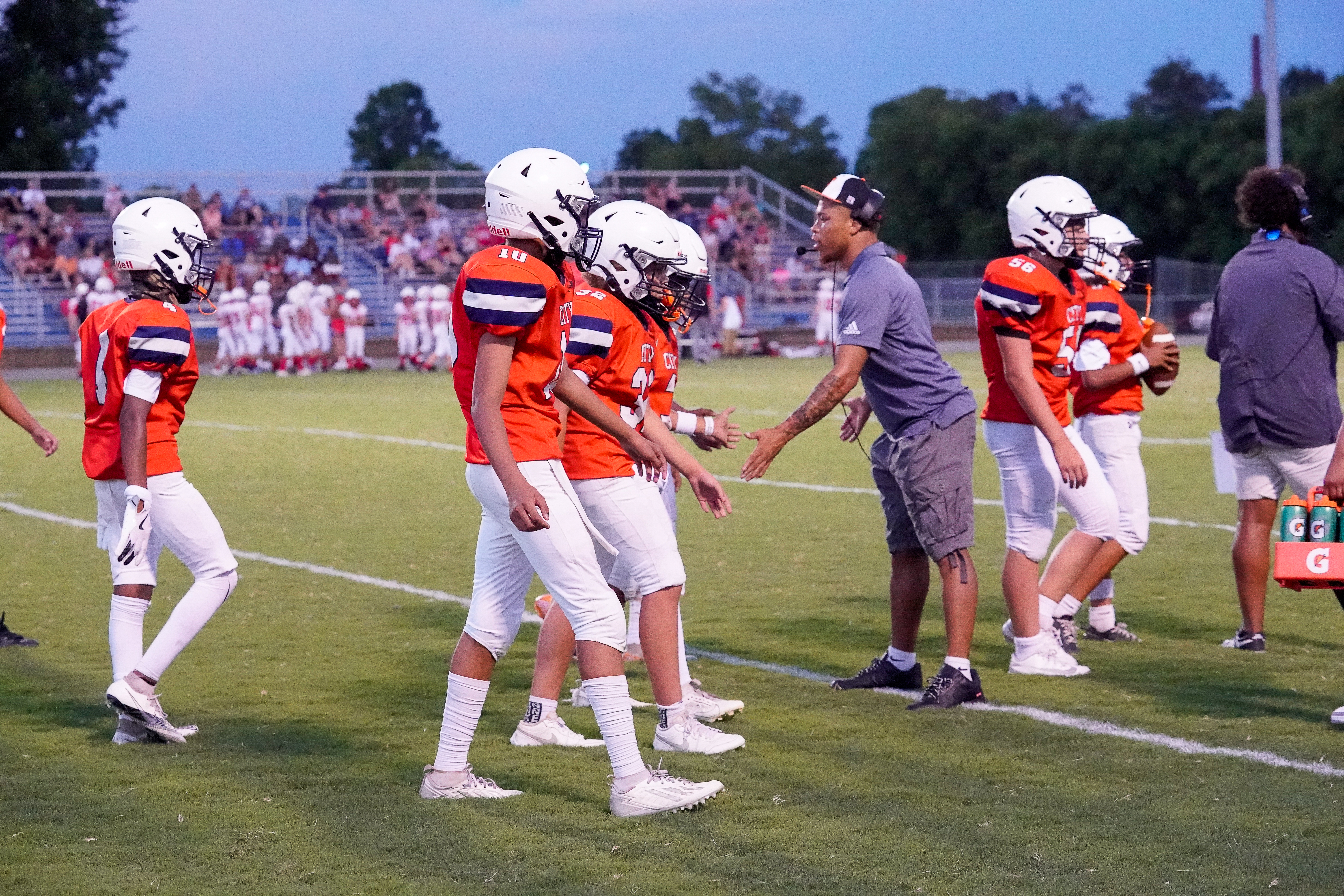 A group of young football players in orange jerseys and white helmets walk across a grassy field towards a coach, with spectators in the background.