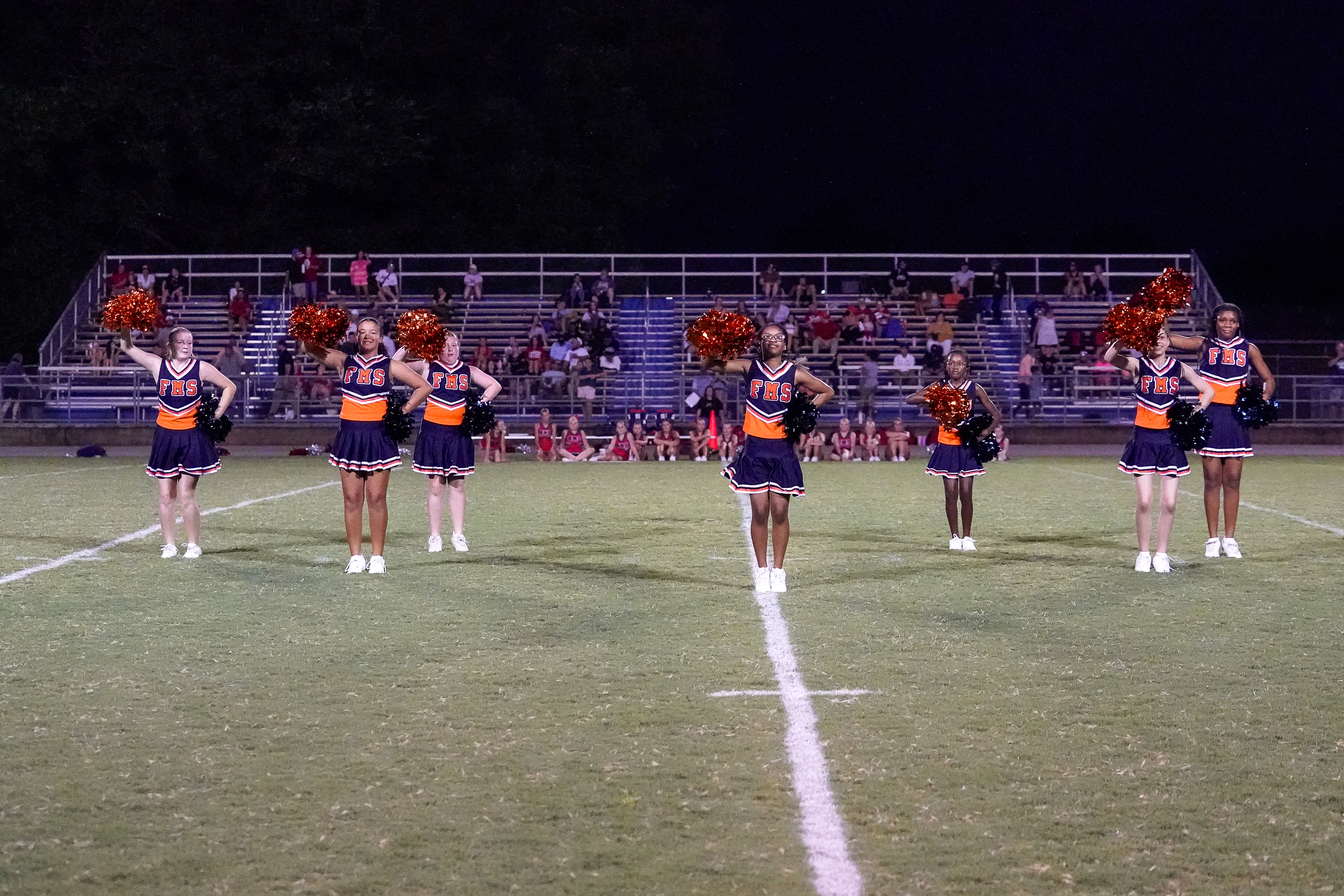 Six cheerleaders in orange and navy uniforms with pom-poms stand on a grassy football field in front of bleachers filled with spectators.