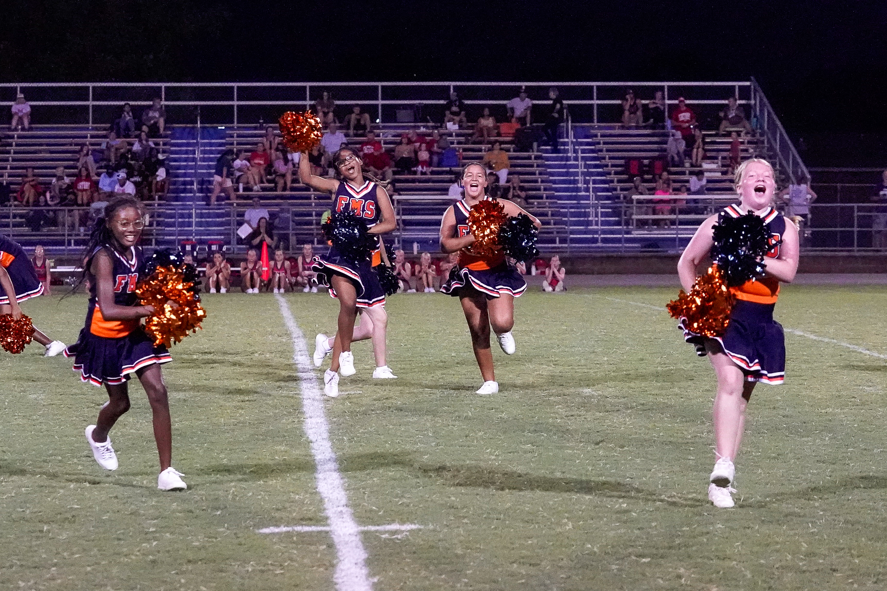 Four cheerleaders in orange and navy uniforms with pom-poms run across a grassy football field in front of a bleacher-filled stadium at night.