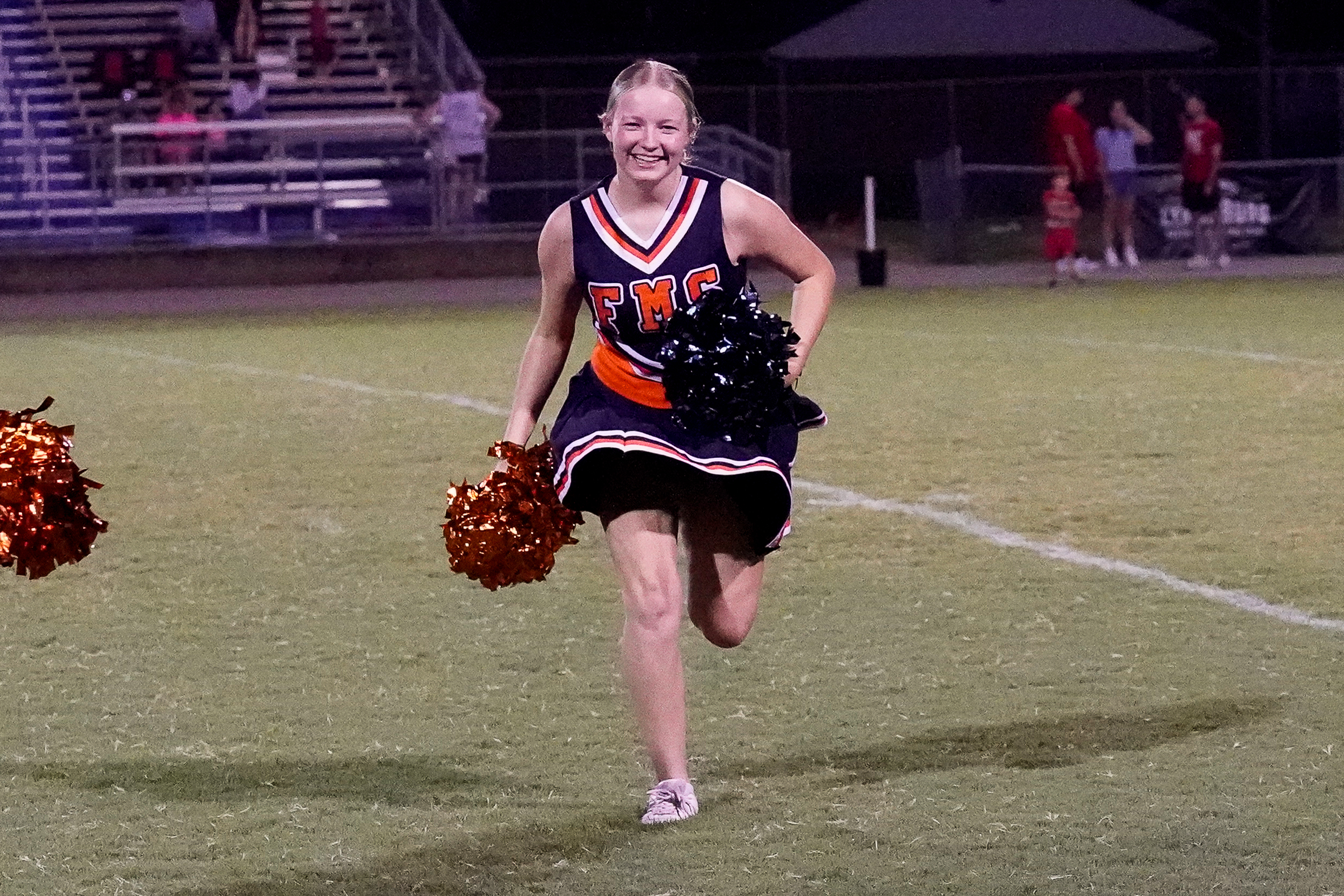 A smiling cheerleader with orange and black pom-poms runs across a grassy field in front of bleachers filled with spectators.