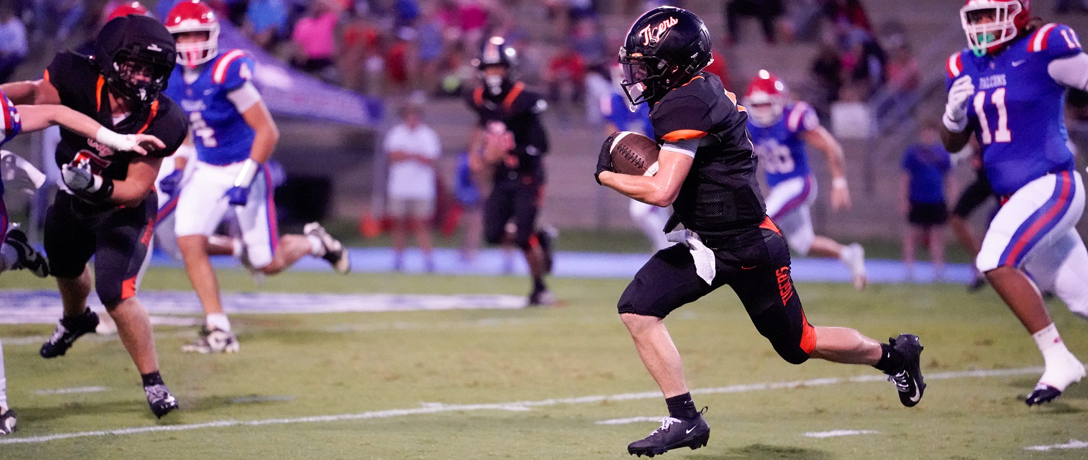 A football player in a black jersey runs with the ball while other players in red, white, and blue uniforms pursue him on a field with spectators in the background.