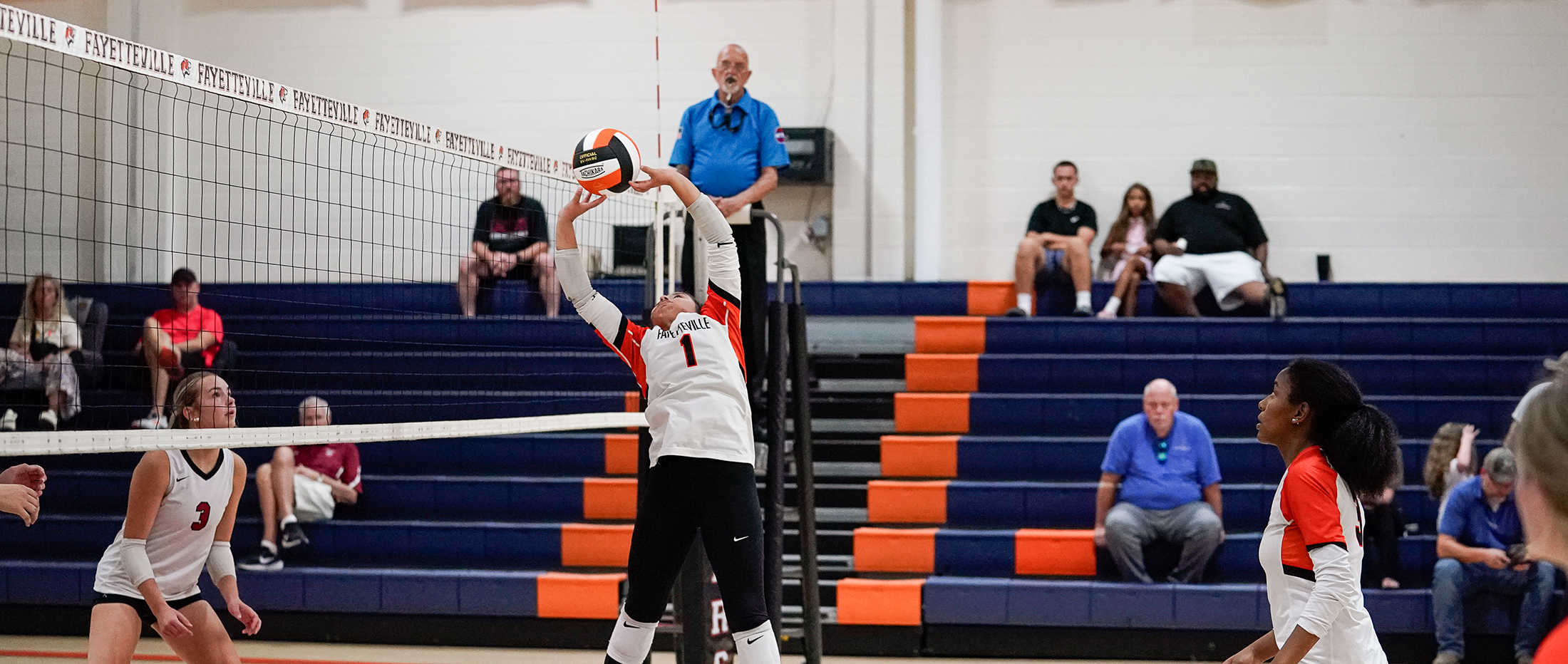 A volleyball player in the foreground is serving the ball over the net, while spectators watch from bleachers in the background.
