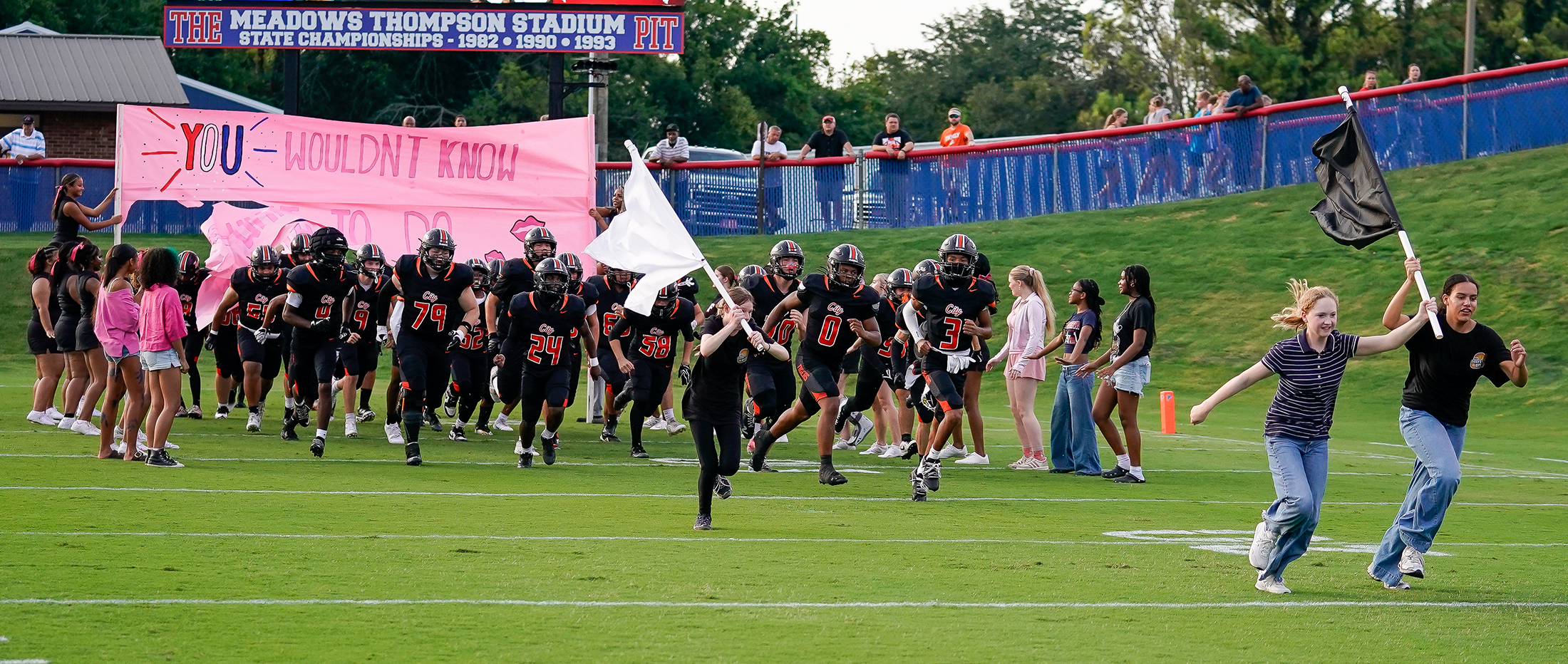 A football team runs onto the field through a pink banner held by supporters, with a stadium and spectators in the background.