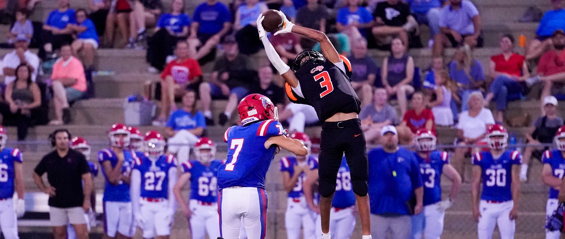 A football player in an orange jersey leaps to catch a pass while a player in a blue jersey defends, with a crowd watching from the bleachers in the background.