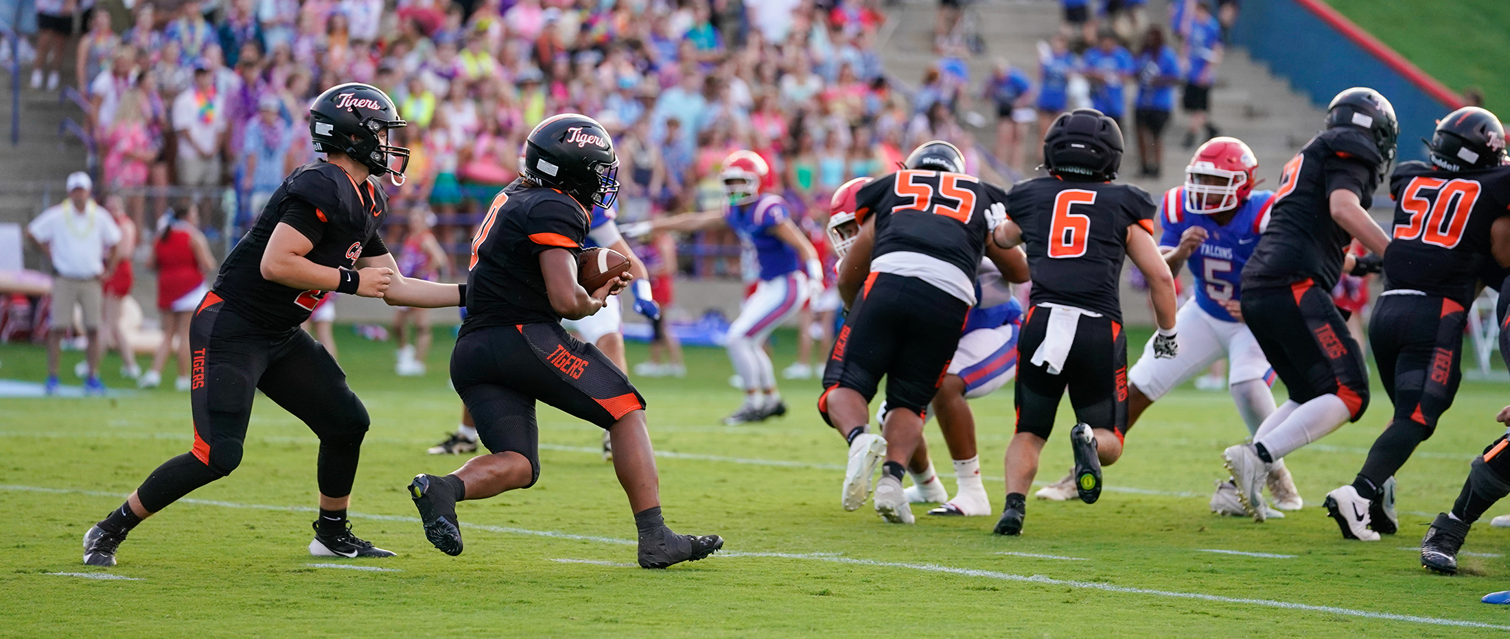 Football players in black and orange uniforms are on the field during a game, with a crowd of spectators in colorful attire in the background.