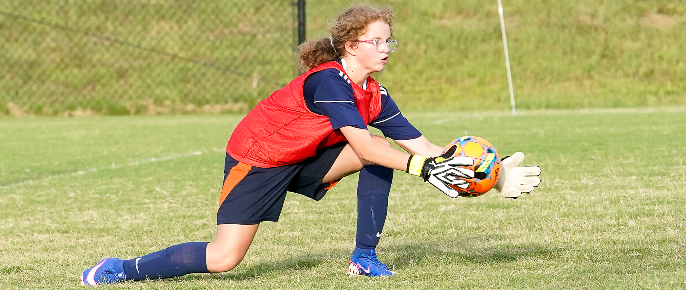 A young soccer goalie in a red vest and blue uniform lunges to catch a ball on a grassy field with a fence in the background.