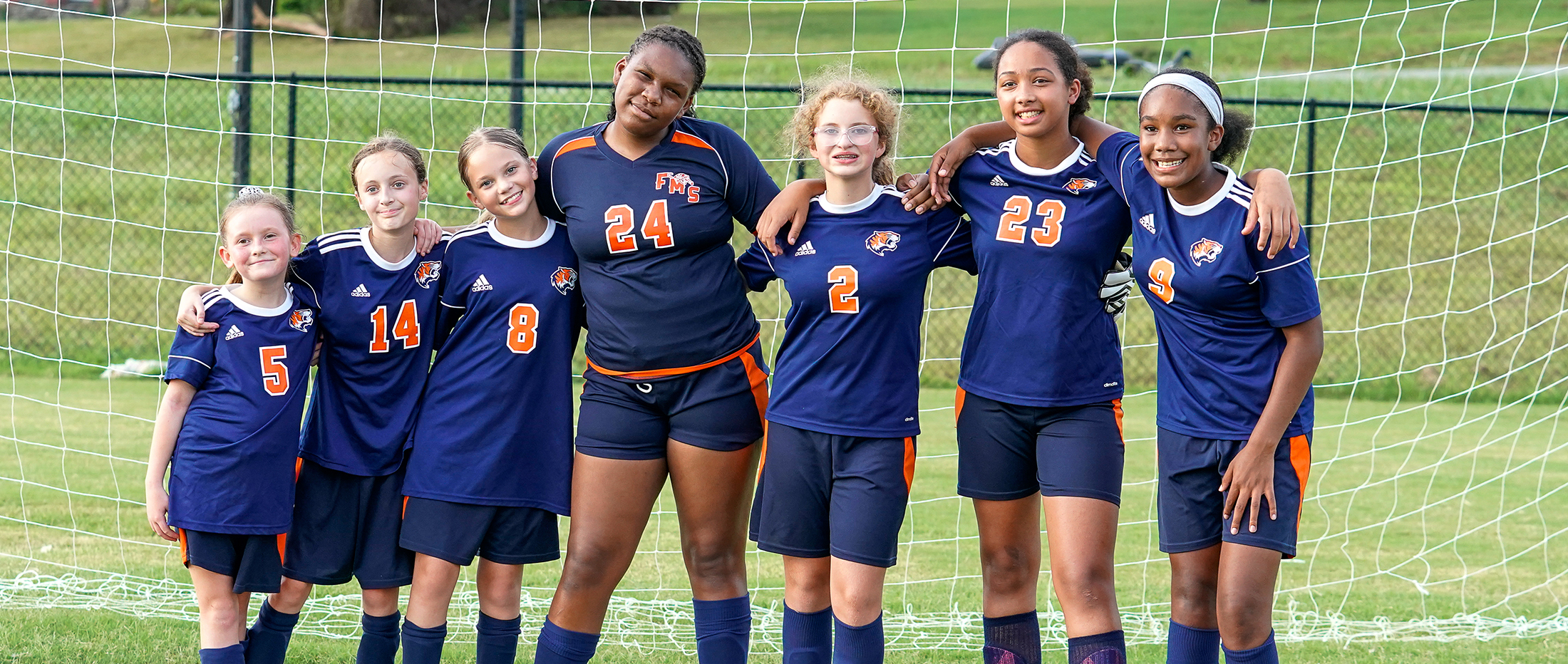 Seven young girls in matching soccer uniforms stand together in front of a soccer net on a grassy field.