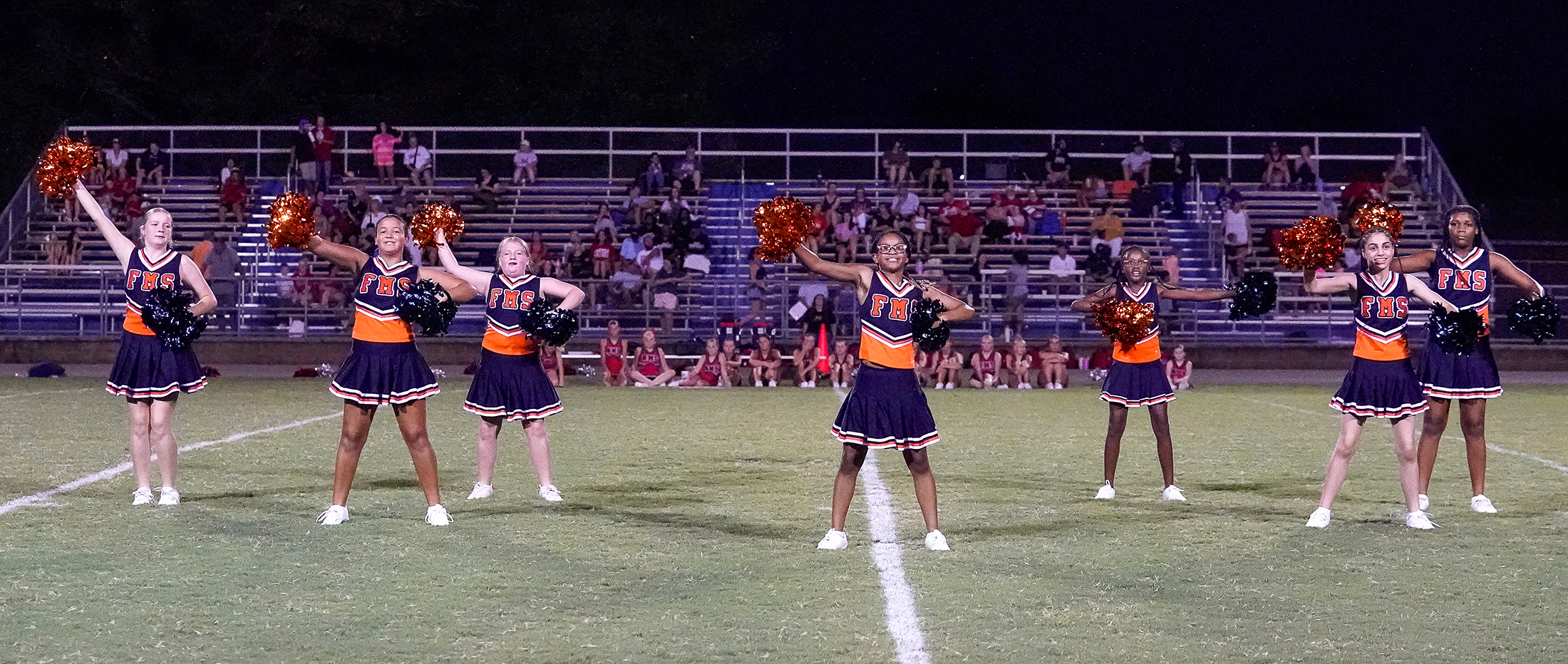 A group of cheerleaders in orange and navy uniforms perform on a grassy field in front of bleachers filled with spectators.