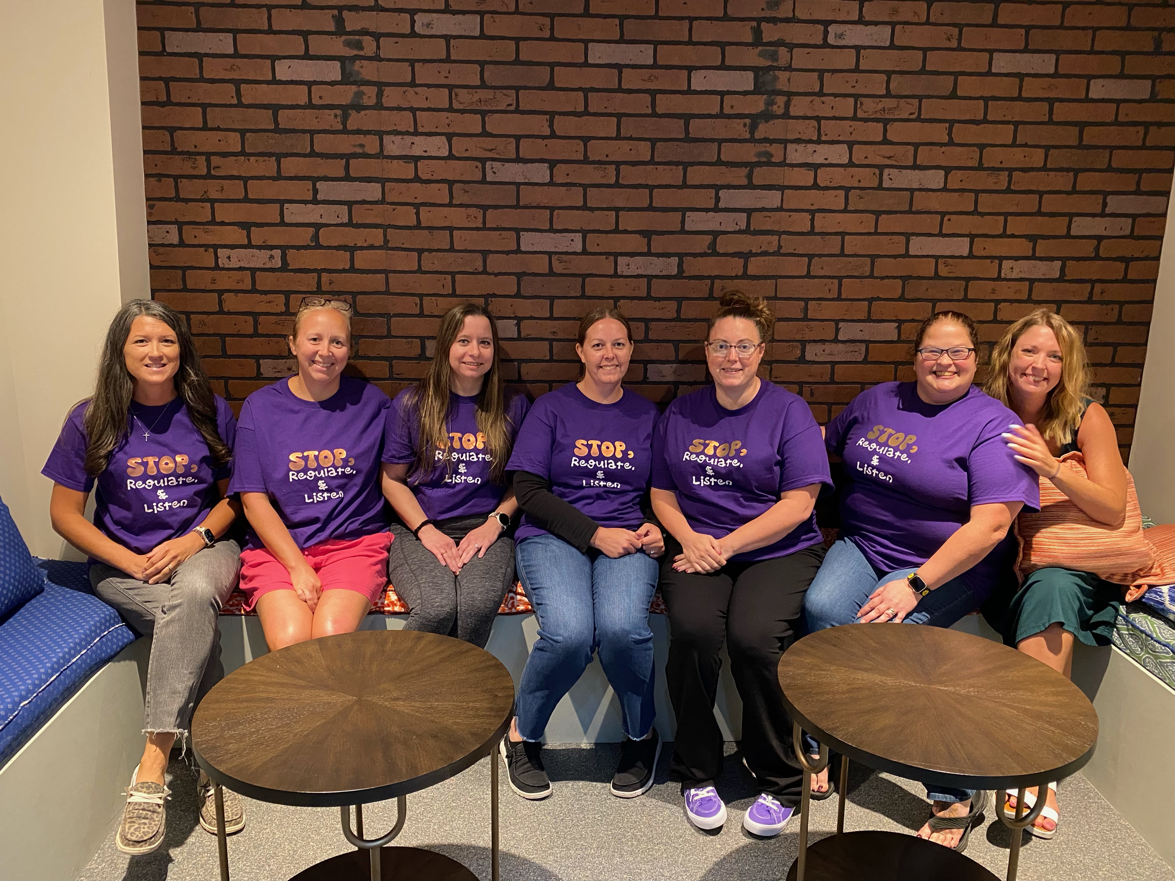 Six women in purple t-shirts sit on a bench in front of a brick wall, with two round tables in the foreground.